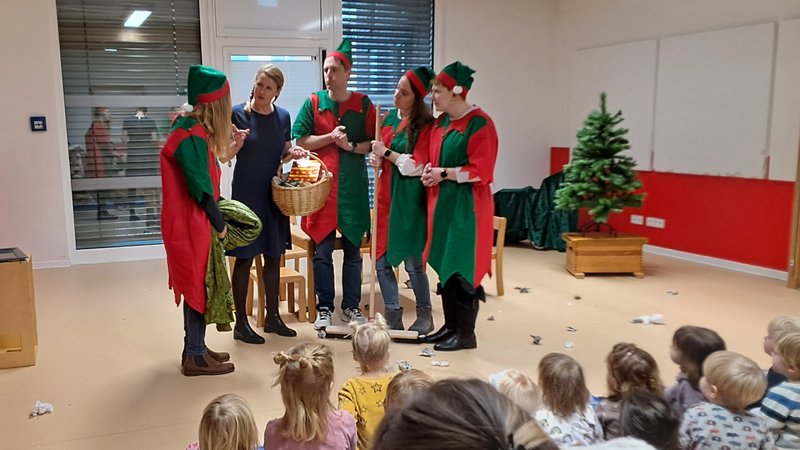Auf dem Bild ist eine Aufführung oder ein kleines Theaterstück in einem Innenraum zu sehen, wahrscheinlich in einer Kindertagesstätte, Schule oder einem Gemeindezentrum, da viele Kinder zuschauen. Hier ist eine detaillierte Beschreibung: Akteure (Erwachsene): In der Mitte stehen vier erwachsene Personen, die in grün-roten Kostümen (ähnlich wie Elfen oder Wichtel) mit passenden Mützen gekleidet sind. Sie scheinen miteinander zu interagieren oder eine Szene aufzuführen. Eine fünfte erwachsene Person in normaler dunkler Kleidung steht ebenfalls bei der Gruppe und hält einen Korb. Publikum (Kinder): Eine Gruppe von Kindern sitzt auf dem Boden und auf Matten im Vordergrund und schaut der Aufführung zu. Man sieht sie hauptsächlich von hinten. Bühne und Requisiten: Die Akteure stehen in einem offenen Bereich. Es gibt einen Korb mit Inhalt (vielleicht Obst oder kleine Geschenke), der als Requisite dient. Dekoration: Rechts im Bild steht ein kleiner, geschmückter Weihnachtsbaum in einem Holztopf, was auf eine Veranstaltung in der Advents- oder Weihnachtszeit hindeutet. Auf dem Boden liegen vereinzelt Papierschnipsel oder Dekorationen. Raum: Der Raum hat helle Wände und einen holzfarbenen Boden. Man sieht große Fenster mit Jalousien und helle Deckenbeleuchtung. Links im Bild steht ein Stativ (wahrscheinlich für eine Kamera oder Videokamera).