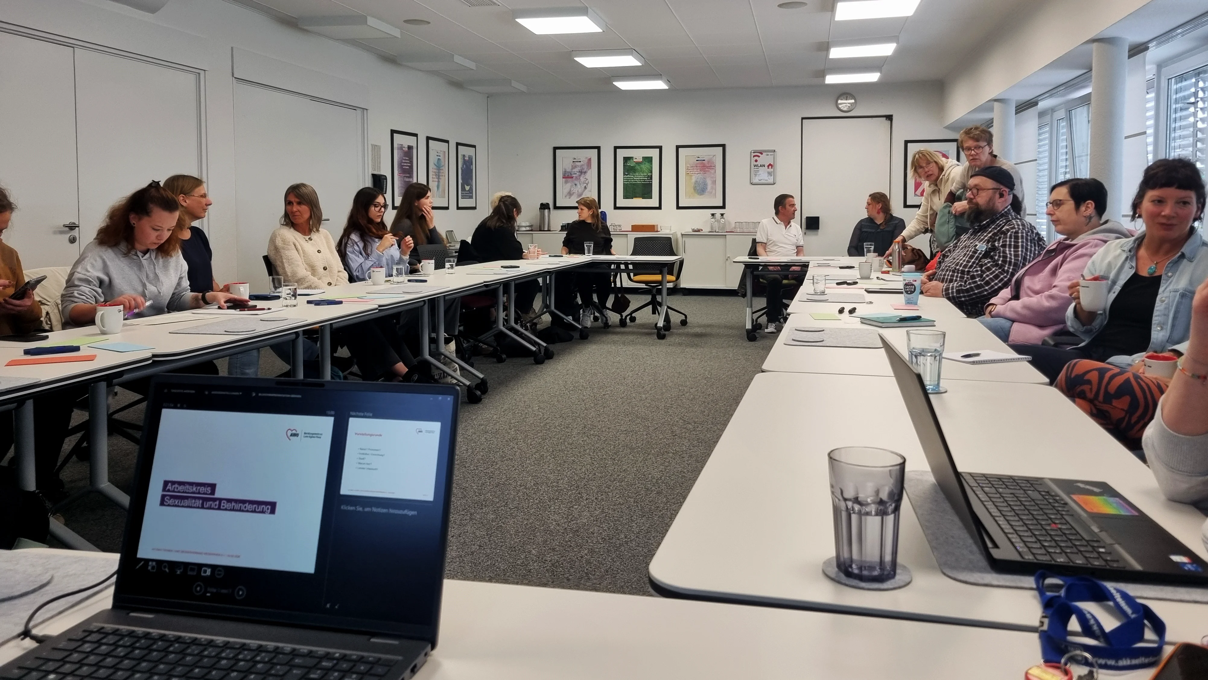 Auf dem Bild sieht man eine größere Gruppe von Menschen in einem Seminar- bzw. Konferenzraum, die an einem U-förmig gestellten Tisch sitzen und an einer Sitzung oder Fortbildung teilnehmen. Auf den Tischen liegen Unterlagen, Notizblöcke, Getränke und Laptops; im Vordergrund ist ein aufgeklappter Laptop zu sehen, auf dessen Präsentationsfolie „Arbeitskreis Sexualität und Behinderung“ steht.