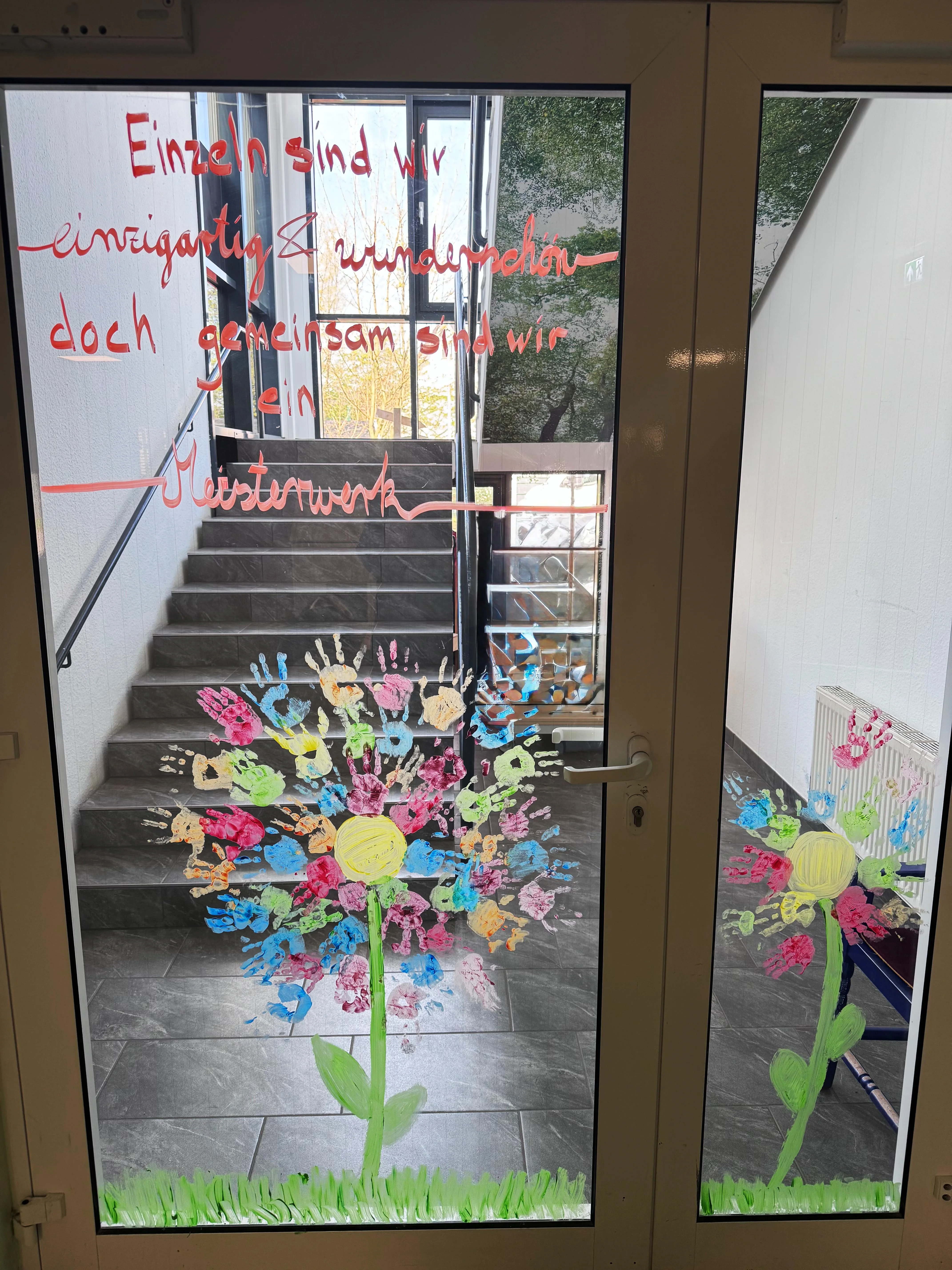 Auf dem Bild ist eine Fenster- bzw. Türgestaltung in einem Gebäude zu sehen. Im Einzelnen: Eine große Blume aus Handabdrücken: Die Blütenblätter bestehen aus vielen bunten Handabdrücken von Kindern in verschiedenen Farben (rot, blau, gelb, orange, pink, grün), die rund um eine gelbe Mitte angeordnet sind. Stiel und Blätter sind grün gemalt, unten gibt es grünes Gras als Abschluss. Ein handgeschriebener Spruch in roter Farbe auf der Glasscheibe: „Einzeln sind wir einzigartig & wunderschön – doch gemeinsam sind wir ein Meisterwerk" Im Hintergrund ist ein Treppenhaus zu sehen, und auf der rechten Seite spiegelt sich eine zweite, kleinere Version der Blume auf einem weiteren Glaselement wider. Das Ganze ist eine wunderschöne, kreative Gemeinschaftsarbeit der Kinder zum Thema Zusammenhalt und Gemeinschaft – perfekt als Dekoration und als Botschaft nach innen und außen.