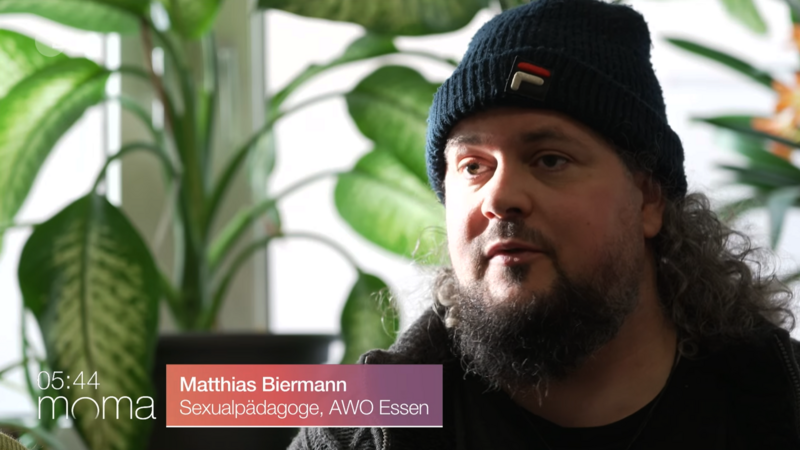 Auf dem Bild ist eine Person in Innenraum-Umgebung vor großen grünen Zimmerpflanzen zu sehen, deren Gesicht absichtlich unkenntlich gemacht wurde.​ Unten links steht eine Einblendung mit der Uhrzeit „05:44“ und dem Schriftzug „moma“, rechts daneben unten ein Namensbalken mit der Beschriftung „Matthias Biermann, Sexualpädagoge, AWO Essen“.