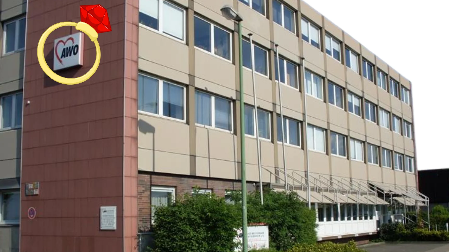 Auf dem Foto ist ein mehrstöckiges Gebäude zu sehen, ein Büro- oder Verwaltungsgebäude. An der Ecke des Gebäudes befindet sich ein Schild mit dem Logo der AWO (Arbeiterwohlfahrt), einer deutschen Wohlfahrtsorganisation. Das Logo besteht aus einem roten Herz mit den Buchstaben "AWO" darin. Auffällig ist, dass auf das Logo nachträglich ein großes Ring-Symbol mit einem roten Edelstein digital eingefügt wurde, das wie ein Verlobungs- oder Schmuckring aussieht. Dieses Symbol wurde als Grafik über das Originalbild gelegt und ist nicht Teil des Gebäudes selbst.
