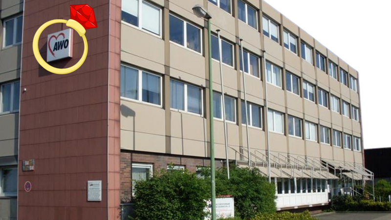 Auf dem Foto ist ein mehrstöckiges Gebäude zu sehen, ein Büro- oder Verwaltungsgebäude. An der Ecke des Gebäudes befindet sich ein Schild mit dem Logo der AWO (Arbeiterwohlfahrt), einer deutschen Wohlfahrtsorganisation. Das Logo besteht aus einem roten Herz mit den Buchstaben "AWO" darin. Auffällig ist, dass auf das Logo nachträglich ein großes Ring-Symbol mit einem roten Edelstein digital eingefügt wurde, das wie ein Verlobungs- oder Schmuckring aussieht. Dieses Symbol wurde als Grafik über das Originalbild gelegt und ist nicht Teil des Gebäudes selbst.
