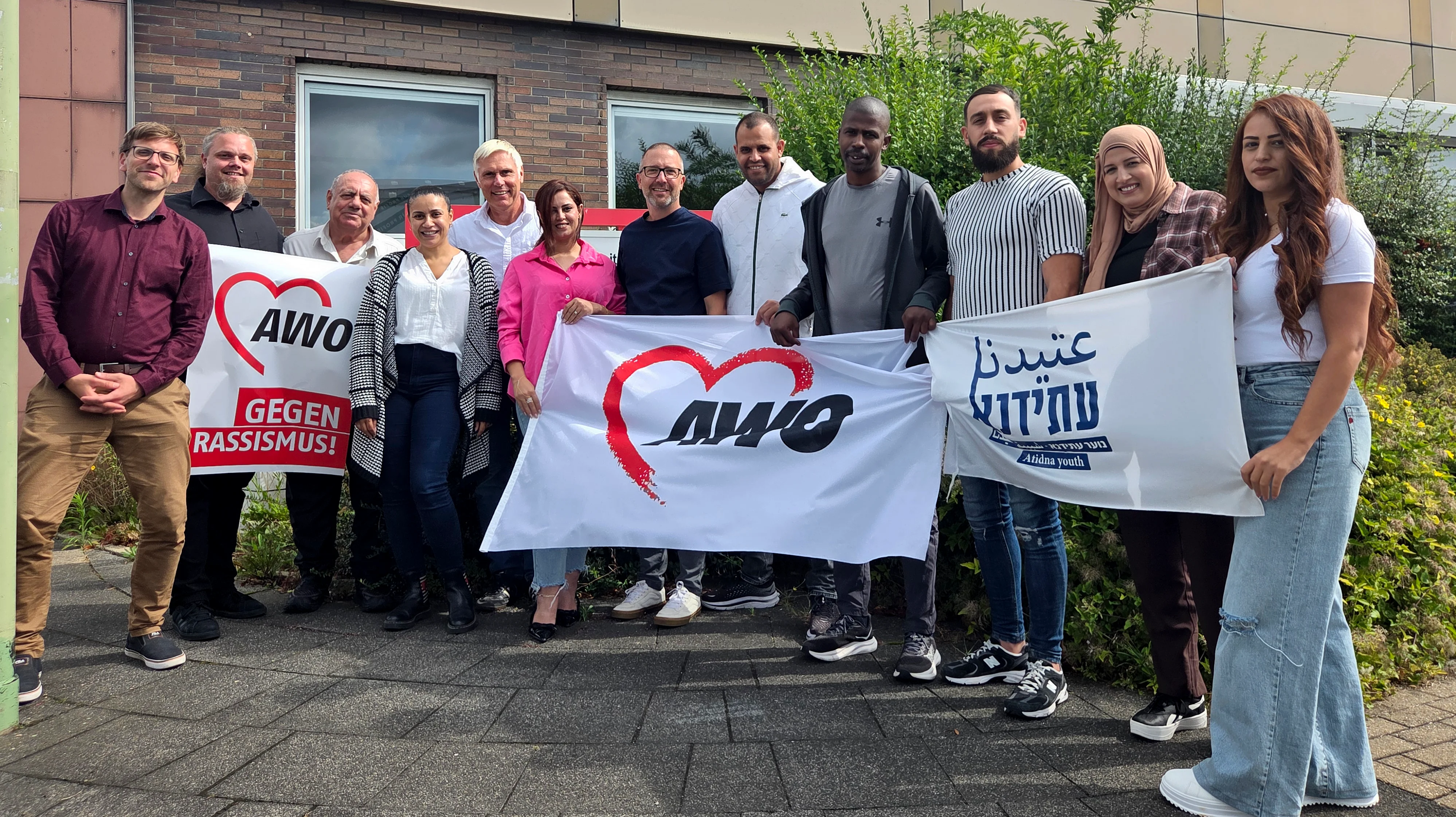 Auf dem Foto ist eine Gruppe von etwa einem Dutzend Personen zu sehen, die vor einem Gebäude stehen. Sie halten drei Banner hoch: Ein weißes Banner mit einem roten Herz und dem Slogan "AWO" in Rot. Ein weiteres weißes Banner mit einem roten Herz und dem Text "AWO GEGEN RASSISMUS!" in Schwarz. Ein weißes Banner mit Text in einer ausländischen Schrift. Die meisten Personen auf dem Foto tragen lässige Kleidung. Die Stimmung wirkt positiv und die Personen schauen freundlich in die Kamera.