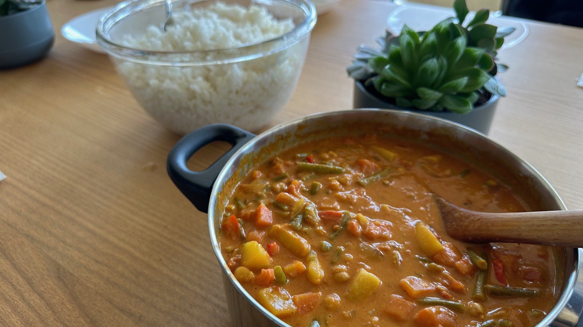 Auf dem Foto ist Folgendes zu sehen: Im Vordergrund: Ein silberner Kochtopf mit schwarzem Griff, gefüllt mit einem bunten Gemüsecurry. Gut erkennbar sind darin: Kartoffelstücke (gelb/cremefarbene Würfel) Möhren (orangefarbene Stücke) Grüne Bohnen Paprika (rote Stücke) Eine cremig-rote Currysauce Ein Holzkochlöffel steckt im Topf Im Hintergrund: Eine transparente Glasschüssel mit weißem Reis (Beilagen) Ein kleiner grüner Topfpflanze (eine Sukkulente oder Kräuterpflanze) Ein heller Holztisch