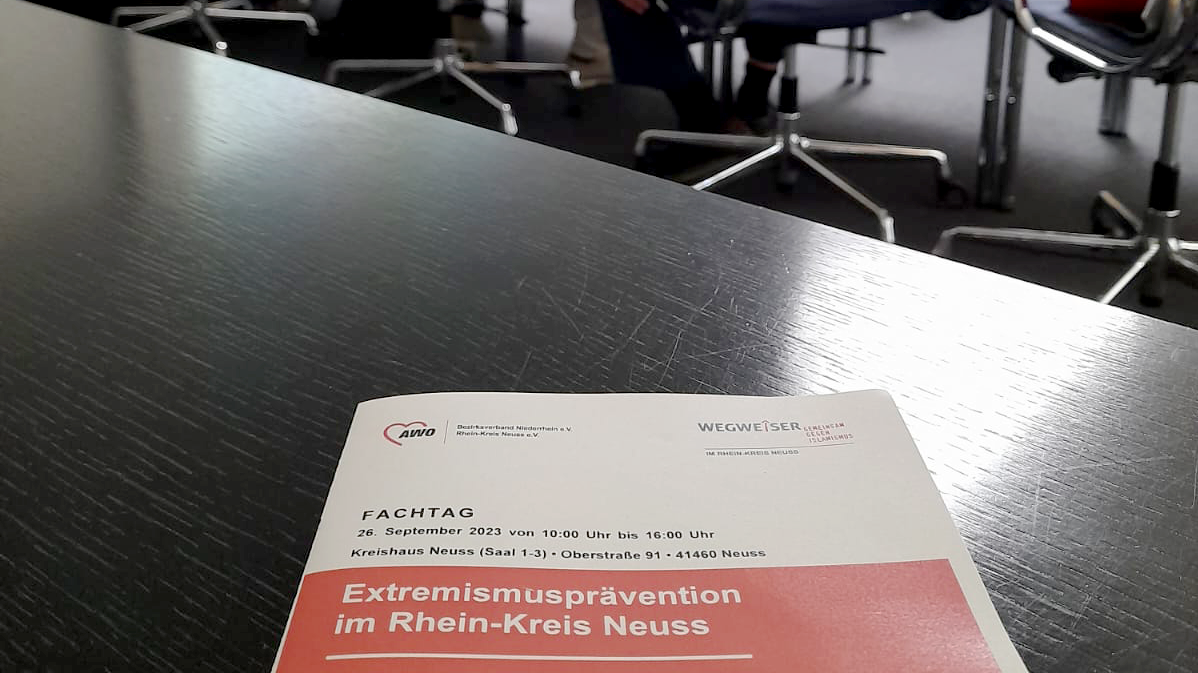 Das Foto zeigt eine Konferenz. Auf dem Flyer im Vordergrund steht: Fachtag September 2023 von 10:00 Uhr bis 16:00 Uhr Kreishaus Neuss (Saal 1-3) - Oberstraße 91 - 41460 Neuss Extremismusprävention im Rhein-Kreis Neuss.