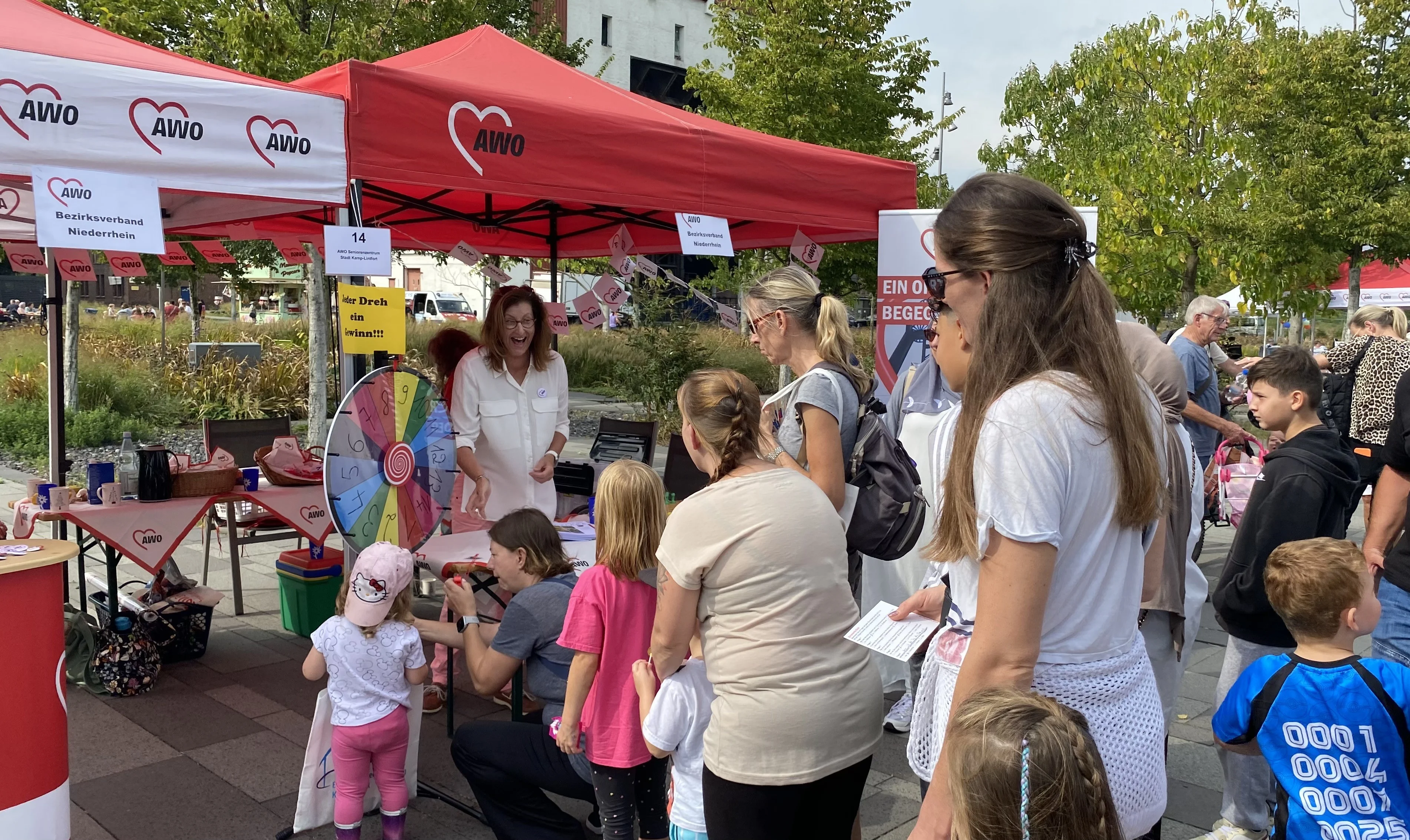 Auf dem Foto ist ein belebter Stand der AWO (Arbeiterwohlfahrt) bei einer öffentlichen Veranstaltung zu sehen. Mehrere Erwachsene und Kinder befinden sich vor den roten AWO-Pavillons. In der Mitte steht eine Frau mit einem Spinnrad, das mit verschiedenen Preisen beschriftet ist und vermutlich für ein Gewinnspiel genutzt wird. Im Hintergrund sind weitere Menschen und Bäume erkennbar. Das Foto fängt eine fröhliche und aktive Szene ein.