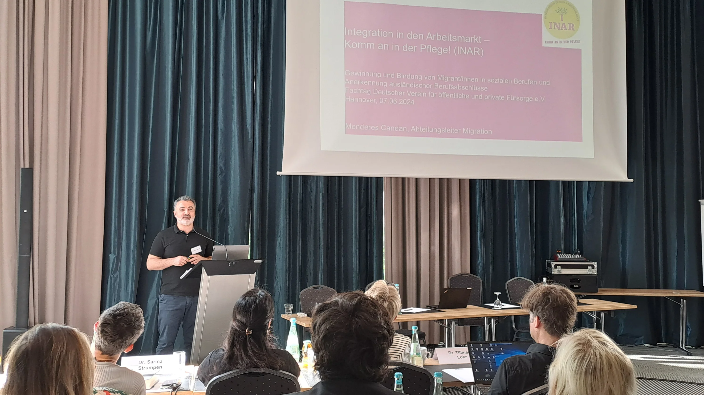 Das Foto zeigt einen Seminarraum, in dem Teilnehmende an Tischen sitzen und auf eine Präsentationswand schauen. Ein Vortragender steht links von der Leinwand, auf der Text und Logos angezeigt werden.