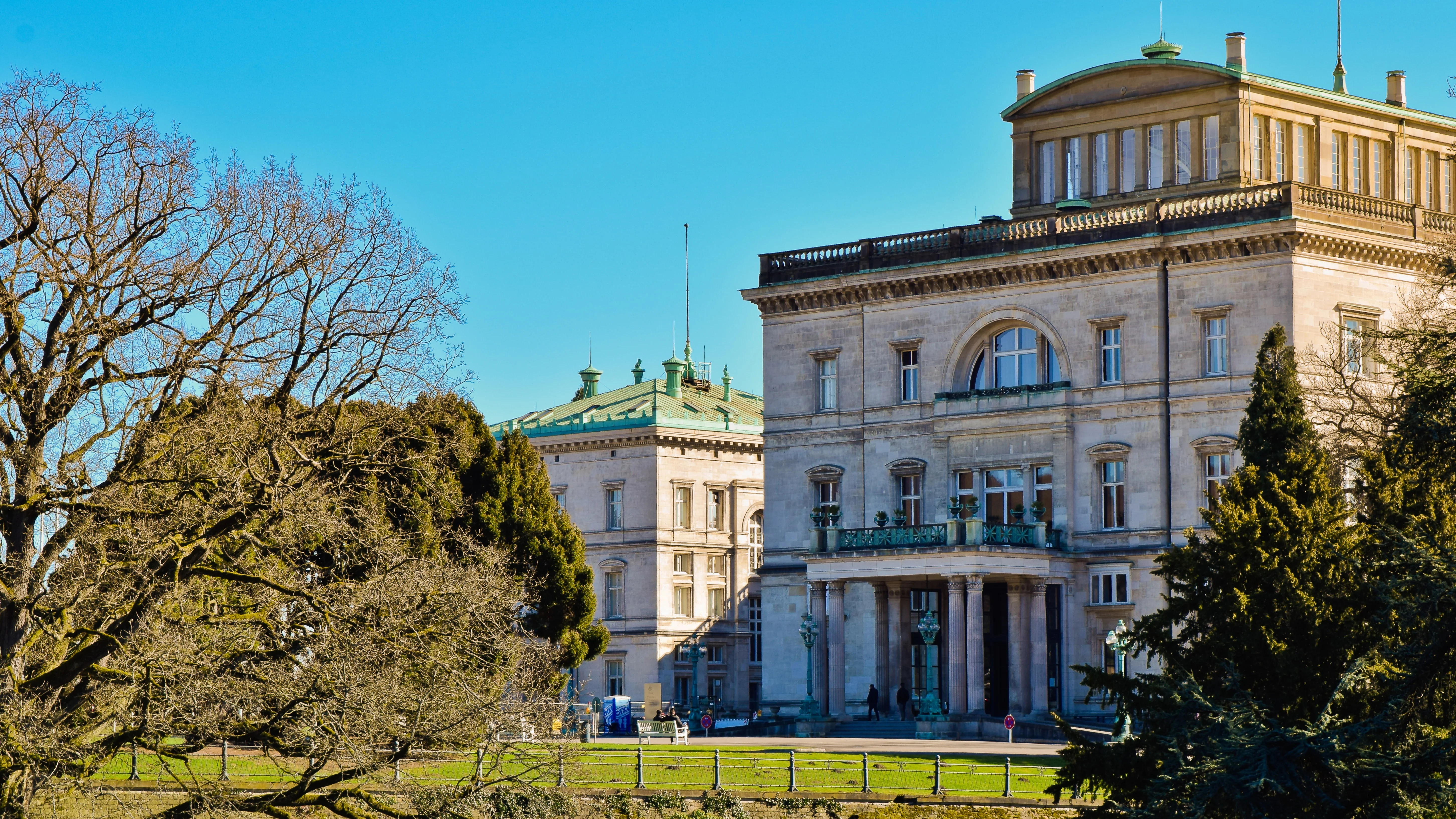 Das Foto zeigt ein imposantes Gebäude mit klassischen architektonischen Merkmalen, darunter Säulen und dekorative Balustraden. Das Gebäude ist von einem gepflegten Rasen und verschiedenen Bäumen umgeben. Einige Bäume sind kahl, während andere immergrün sind, was darauf hindeutet, dass das Foto möglicherweise in einer Jahreszeit aufgenommen wurde, in der einige Bäume ruhen. Der Himmel ist klar und blau, was auf einen sonnigen Tag hindeutet.