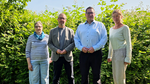 Auf dem Bild stehen vier Personen im Freien vor einer dichten, grünen Hecke bzw. Buschreihe auf einer Wiese. Alle stehen in einer Reihe und schauen zur Kamera, drei tragen lange Hosen und Oberteile in eher hellen, neutralen Farben, eine Person links ein gestreiftes Oberteil mit blauem Halstuch.