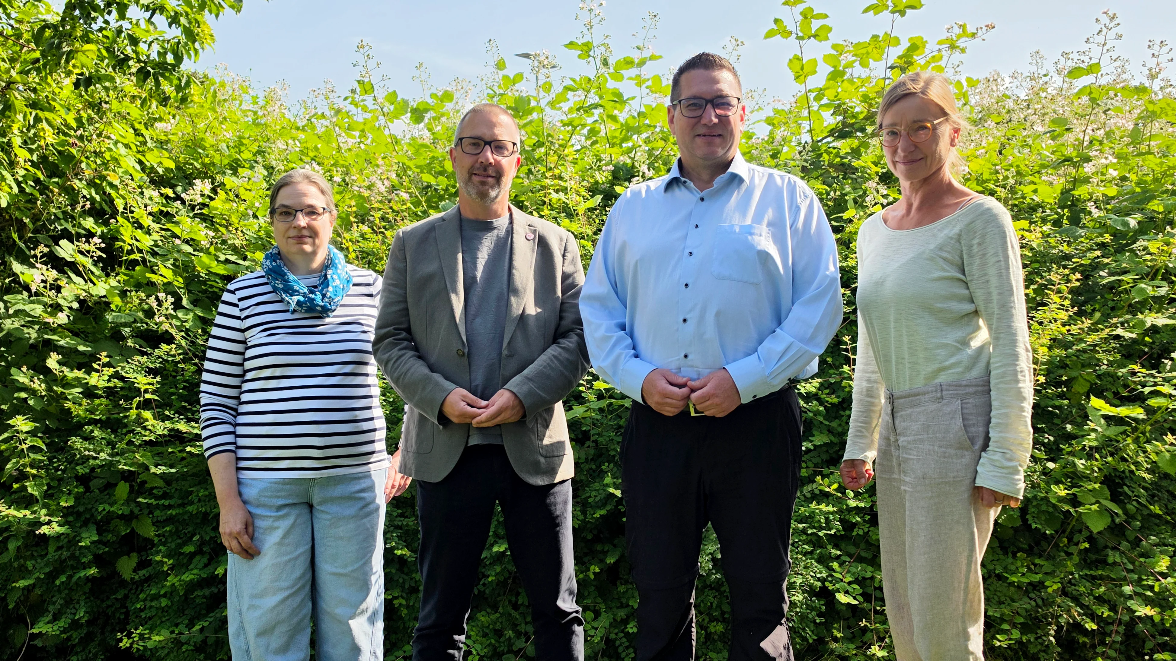 Auf dem Bild stehen vier Personen im Freien vor einer dichten, grünen Hecke bzw. Buschreihe auf einer Wiese. Alle stehen in einer Reihe und schauen zur Kamera, drei tragen lange Hosen und Oberteile in eher hellen, neutralen Farben, eine Person links ein gestreiftes Oberteil mit blauem Halstuch.