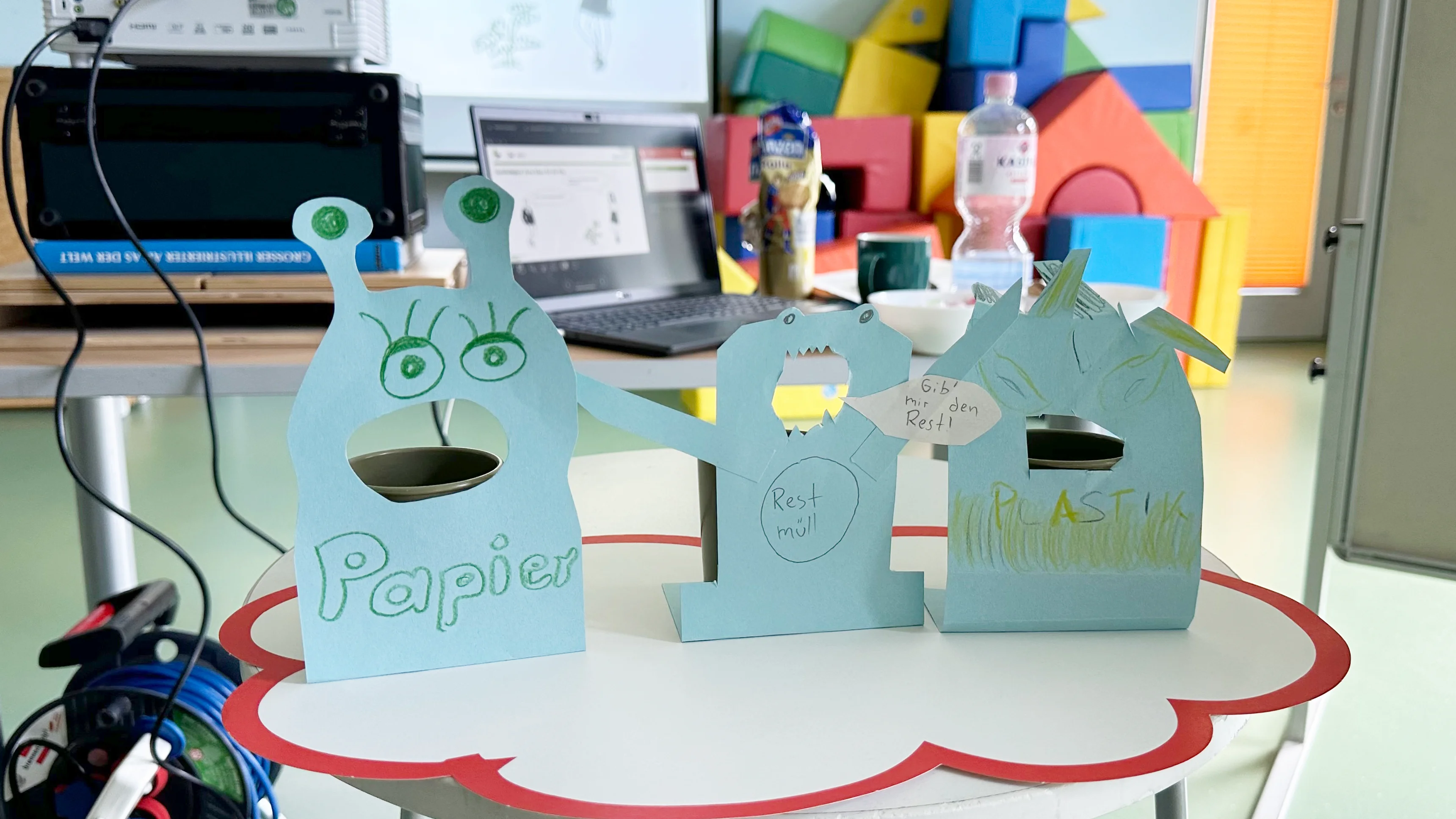 Vorne auf dem Bild stehen drei gebastelte „Müllmonster“ aus hellblauem Papier auf einem runden Tisch mit roter Umrandung. Links ist ein Monster mit Augenfühlern, großem ausgeschnittenen Mund und der Aufschrift „Papier“. In der Mitte steht ein monsterartiger Aufsteller mit geöffnetem Maul und der Beschriftung „Restmüll“, daneben eine Sprechblase „Gib mir den Rest!“. Rechts ist ein weiteres Monster mit Stacheln oder Haaren, vor dem „Plastik“ (bzw. „Plastik/Plastikmüll“) steht. Im Hintergrund sieht man einen Laptop, einen Beamer und bunte Schaumstoff-Bausteine
