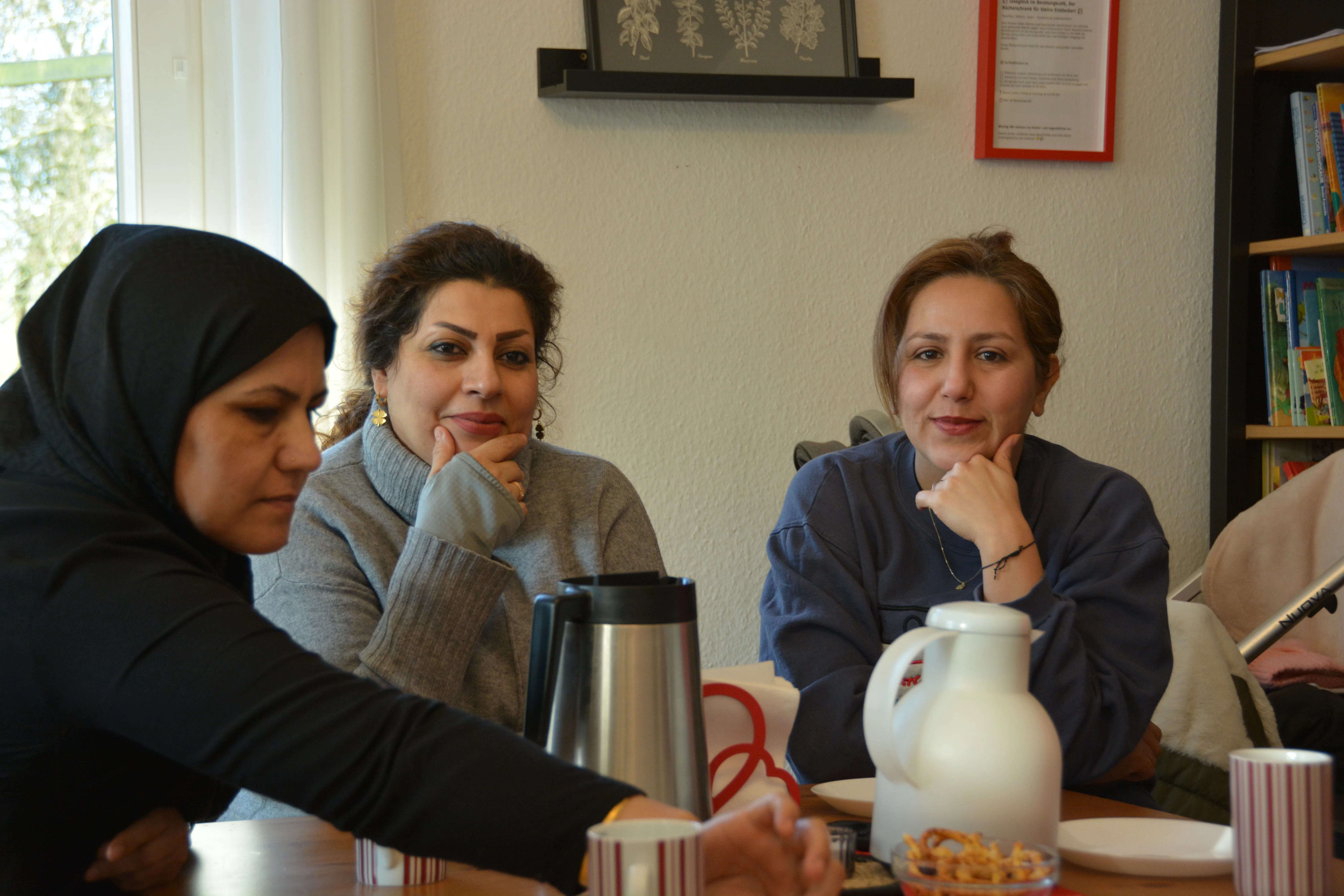 Auf dem Foto, das Sie mir zusammen mit der Frage geschickt haben, sind drei Frauen zu sehen, die an einem Tisch sitzen und sich zu unterhalten scheinen. Die Szene wirkt wie ein informelles Treffen oder Gespräch: Links im Bild sitzt eine Frau mit schwarzem Kopftuch (Hijab) In der Mitte sitzt eine Frau mit dunklen, hochgesteckten Haaren und blauen Ohrringen, die einen grauen Pullover trägt Rechts sitzt eine Frau mit hellbraunen Haaren in einem dunkelblauen Pullover Auf dem Tisch befinden sich eine Thermoskanne, eine weiße Kaffeekanne, Tassen und eine Schale mit Snacks. Im Hintergrund sind Bilderrahmen an der Wand und ein Bücherregal zu sehen. Die Atmosphäre wirkt gemütlich und wie ein persönliches Gespräch oder Meeting in einem Beratungs- oder Büroraum.