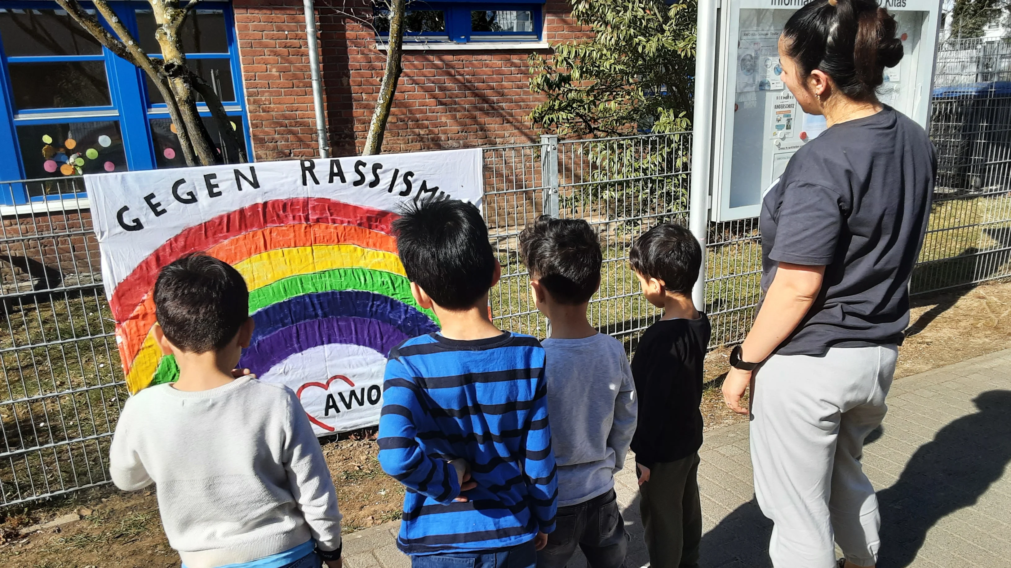 Auf dem Foto ist eine Gruppe von vier Kindern und einer erwachsenen Person zu sehen, die vor einem Zaun stehen und ein großes, selbstgestaltetes Banner betrachten. Das Banner zeigt einen bunten Regenbogen mit der Aufschrift „GEGEN RASSISMUS“ und das Logo der AWO (Arbeiterwohlfahrt). Die Kinder stehen mit dem Rücken zur Kamera, tragen verschiedene legere Kleidung und scheinen sich das Banner gemeinsam anzuschauen. Die erwachsene Person (vermutlich eine Erzieherin oder Betreuerin) steht rechts daneben, ebenfalls mit dem Rücken zur Kamera, und beobachtet die Szene. Im Hintergrund sind ein Gebäude mit blauen Fensterrahmen, ein Baum und ein Infokasten zu sehen. Das Bild vermittelt eine starke Botschaft gegen Rassismus und zeigt eine pädagogische Situation, vermutlich in einer Kita oder Schule, in der Kinder für Vielfalt und Toleranz sensibilisiert werden.