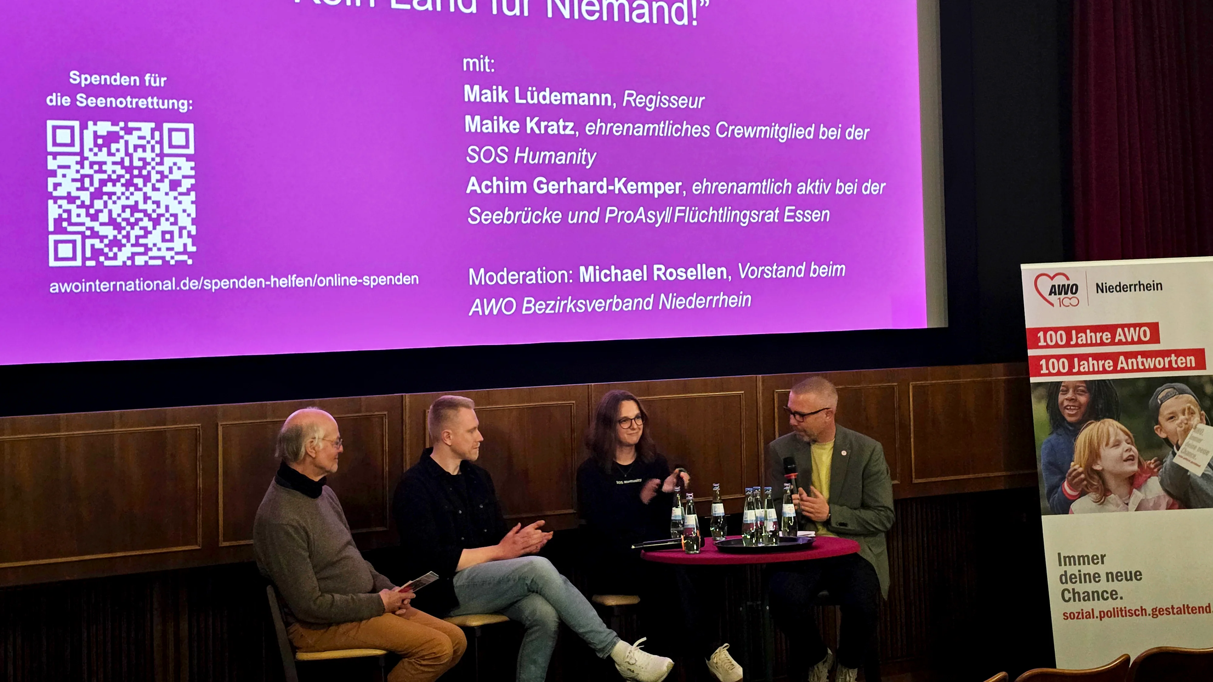 Auf dem Bild ist ein Podiumsgespräch in einem Kinosaal zu sehen. Auf der großen Leinwand steht die Veranstaltung „Publikumsgespräch: ‘Kein Land für Niemand!’“ mit Logos der AWO (Bezirksverband Niederrhein und AWO Essen). Links auf der Leinwand wird zu Spenden für die Seenotrettung aufgerufen, inklusive QR-Code und URL zu awointernational.de. Darunter sind die Namen der Diskutierenden aufgeführt: Maik Lüdemann (Regisseur), Maike Kratz (ehemaliges ehrenamtliches Crewmitglied bei SOS Humanity) und Achim Gerhard-Kemper (ehrenamtlich aktiv bei Seebrücke und ProAsyl Flüchtlingsrat Essen). Im unteren Teil des Bildes sitzen diese drei Personen zusammen mit dem Moderator Michael Rosellen auf einer kleinen Bühne an einem runden Tisch mit Mikrofonen und Wasserflaschen. Rechts steht ein AWO-Roll‑Up mit dem Slogan „Immer deine neue Chance. sozial.politisch.gestaltend.“ und dem Hinweis auf „100 Jahre AWO – 100 Jahre Antworten“.
