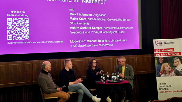 Auf dem Bild ist ein Podiumsgespräch in einem Kinosaal zu sehen. Auf der großen Leinwand steht die Veranstaltung „Publikumsgespräch: ‘Kein Land für Niemand!’“ mit Logos der AWO (Bezirksverband Niederrhein und AWO Essen). Links auf der Leinwand wird zu Spenden für die Seenotrettung aufgerufen, inklusive QR-Code und URL zu awointernational.de. Darunter sind die Namen der Diskutierenden aufgeführt: Maik Lüdemann (Regisseur), Maike Kratz (ehemaliges ehrenamtliches Crewmitglied bei SOS Humanity) und Achim Gerhard-Kemper (ehrenamtlich aktiv bei Seebrücke und ProAsyl Flüchtlingsrat Essen). Im unteren Teil des Bildes sitzen diese drei Personen zusammen mit dem Moderator Michael Rosellen auf einer kleinen Bühne an einem runden Tisch mit Mikrofonen und Wasserflaschen. Rechts steht ein AWO-Roll‑Up mit dem Slogan „Immer deine neue Chance. sozial.politisch.gestaltend.“ und dem Hinweis auf „100 Jahre AWO – 100 Jahre Antworten“.