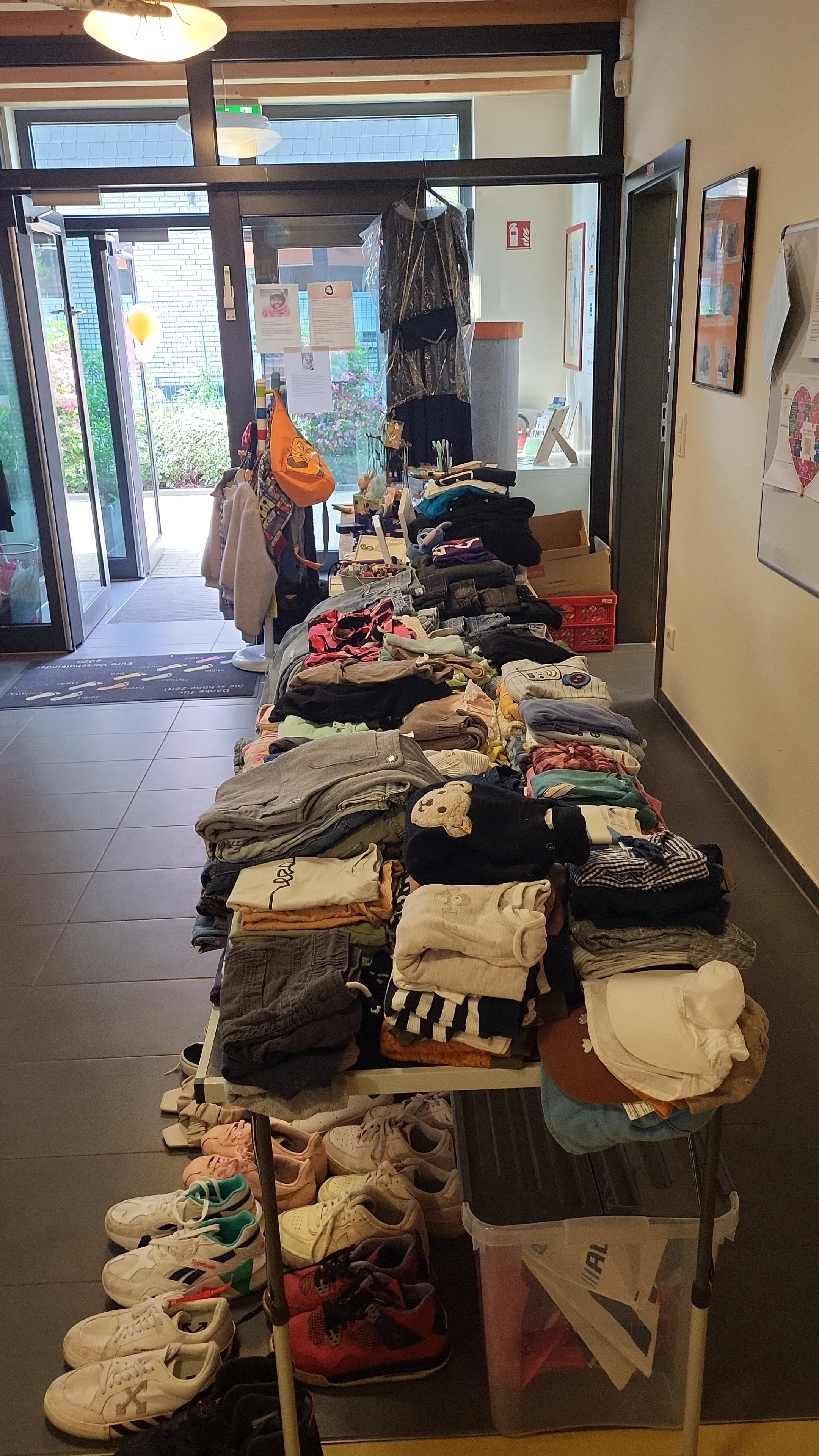 Auf dem Bild sieht man den Eingangs‑ oder Flurbereich eines Gebäudes, in dem auf mehreren Tischen und darunter Kleidung und Schuhe ausgelegt sind. Es wirkt wie eine kleine Kleiderbörse oder Spenden‑/Tauschaktion, vor allem mit gefalteten Kinder‑ und Erwachsenensachen, Schuhpaaren am Boden sowie einigen hängenden Kleidungsstücken und Infoplakaten an den Türen und Wänden.