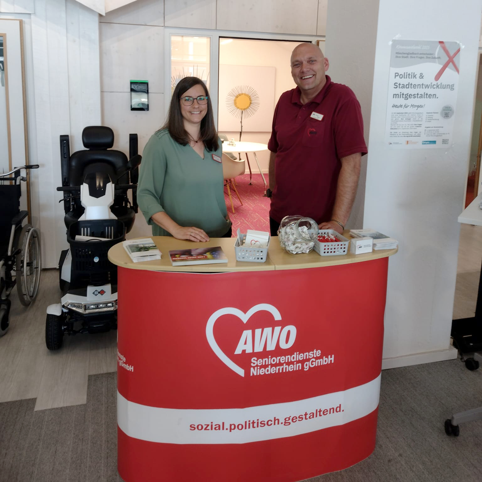Auf dem Foto sind zwei Personen (eine Frau und ein Mann) an einem Infostand der AWO Seniorendienste Niederrhein gGmbH zu sehen. Sie stehen hinter einem rot-weißen Empfangstresen mit dem AWO-Logo und dem Slogan „sozial.politisch.gestaltend.“. Auf dem Tresen liegen einige Informationsmaterialien, Flyer, Broschüren sowie ein Glas mit Bonbons oder ähnlichem. Im Hintergrund sieht man ein kleines, hausähnliches Modell mit einem Fenster, durch das ein Zimmer mit einem sonnendekorierten Spiegel sichtbar ist. Links im Bild sind ein Rollstuhl und ein elektrischer Rollator zu sehen, was auf eine Einrichtung oder Veranstaltung mit Bezug zur Altenhilfe oder Pflege schließen lässt. Rechts an der Wand hängt ein Plakat mit dem Titel „Politik & Stadtentwicklung mitgestalten“, das auf eine Beteiligungsmöglichkeit hinweist – möglicherweise im Rahmen eines Aktionstags oder einer Informationsveranstaltung. Die Atmosphäre wirkt freundlich und einladend – wahrscheinlich handelt es sich um einen Tag der offenen Tür, einen Informationstag oder eine Messe rund um Seniorenarbeit oder Pflege.