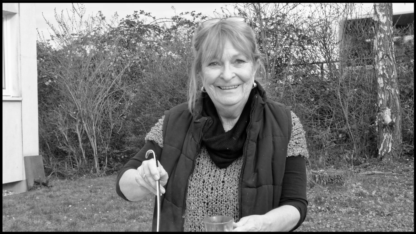Das Foto zeigt eine lächelnde Frau in einem Außenbereich. Sie trägt eine ärmellose Weste über einem gestrickten Pullover und hat ein Getränk in der Hand. Die Frau hat einen Löffel in der Hand. Im Hintergrund sind Büsche und Bäume zu sehen. Das Bild ist in Schwarz-Weiß aufgenommen.