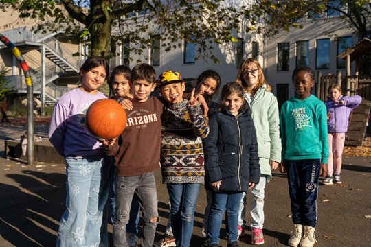 Auf dem Foto ist eine Gruppe von Kindern auf einem Schulhof oder Spielplatz zu sehen. Sie stehen eng beisammen und lächeln in die Kamera, einige legen ihre Arme umeinander oder machen Handzeichen. Ein Kind hält einen orangefarbenen Basketball. Die Kinder sind unterschiedlich gekleidet, einige tragen warme Jacken und andere leichtere Kleidung, was auf eine herbstliche Jahreszeit hindeuten könnte. Im Hintergrund sieht man eine Schule oder ein Gebäude mit großen Fenstern, eine Treppe sowie ein Spielgerüst. Die Sonne scheint, wodurch lange Schatten auf dem Boden entstehen.