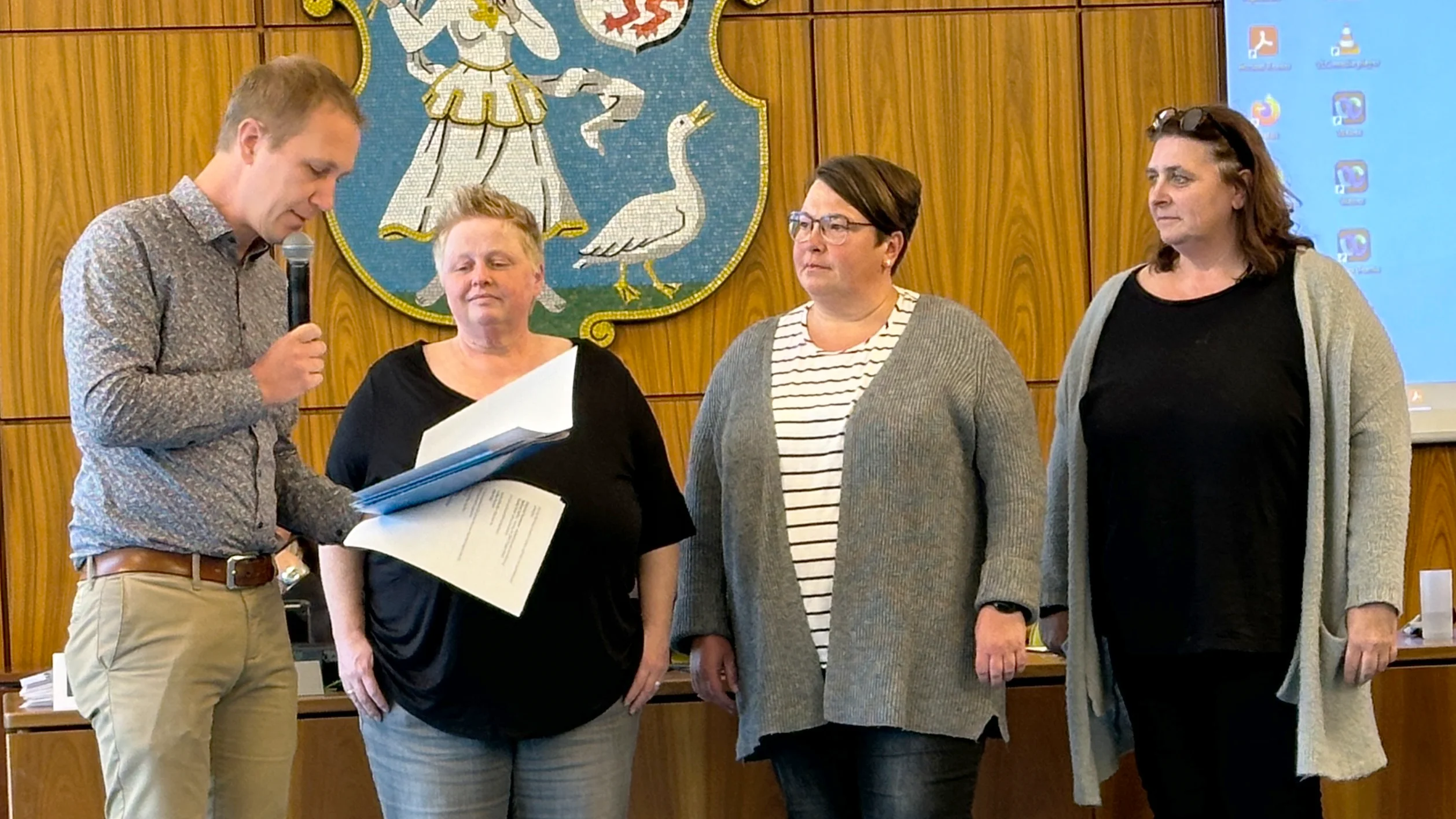 Das Foto zeigt vier Personen, die vor einer Wand mit einem Wappen stehen. Das Wappen hat einen Schild mit einem Löwen und einer Burg. Die Person links hält einen geöffneten Ordner oder ein Dokument, während die anderen neben ihr stehen und vermutlich in Richtung Publikum oder Kamera blicken.