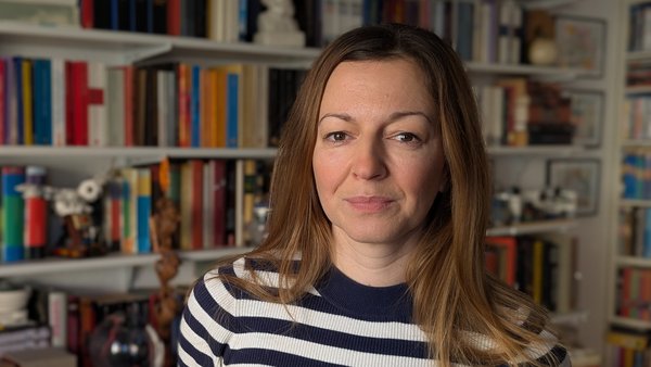 Auf dem Foto ist eine Person mit schulterlangem, hellbraunem Haar in einem dunkelblau‑weiß geringelten Pullover zu sehen. Im Hintergrund steht ein großes Bücherregal mit vielen verschiedenfarbigen Büchern und einigen weiteren Gegenständen wie Kisten, Dekofiguren und Schalen.