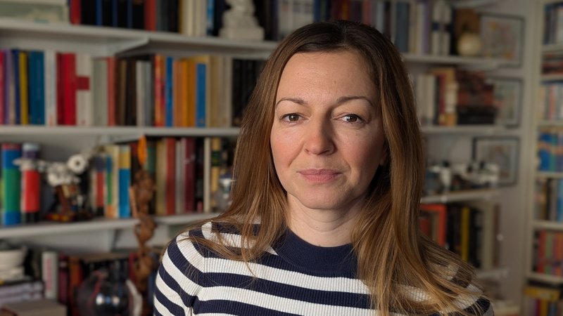 Auf dem Foto ist eine Person mit schulterlangem, hellbraunem Haar in einem dunkelblau‑weiß geringelten Pullover zu sehen. Im Hintergrund steht ein großes Bücherregal mit vielen verschiedenfarbigen Büchern und einigen weiteren Gegenständen wie Kisten, Dekofiguren und Schalen.