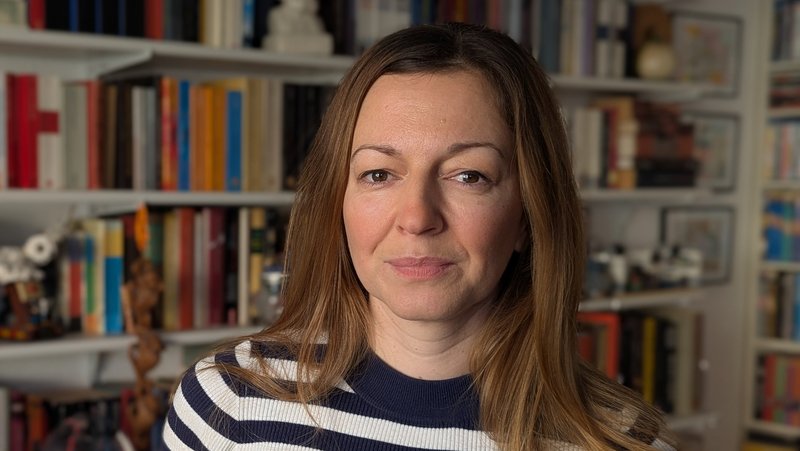 Auf dem Foto ist eine Person mit schulterlangem, hellbraunem Haar in einem dunkelblau‑weiß geringelten Pullover zu sehen. Im Hintergrund steht ein großes Bücherregal mit vielen verschiedenfarbigen Büchern und einigen weiteren Gegenständen wie Kisten, Dekofiguren und Schalen.