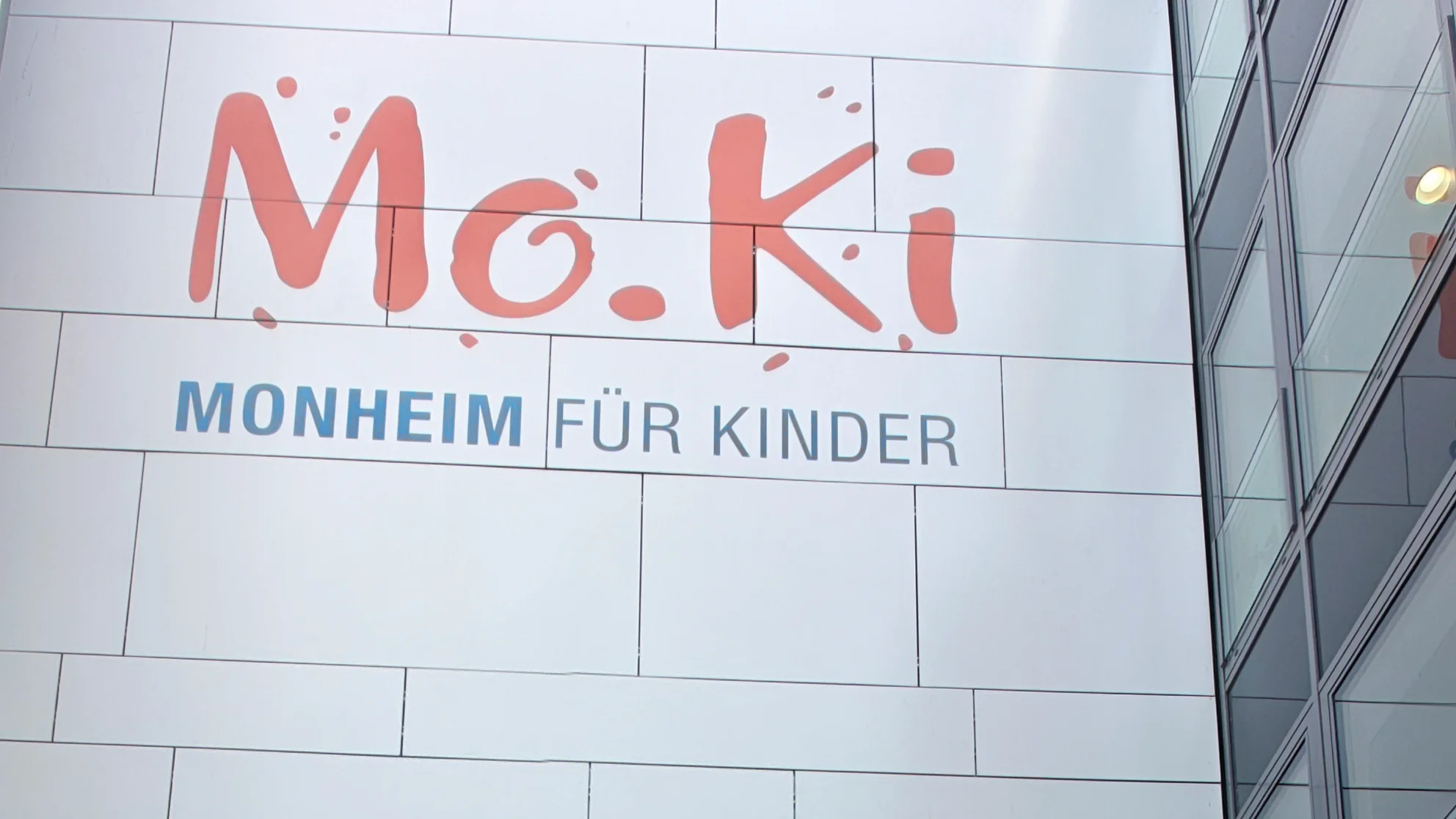 Man sieht man die Fassade eines modernen Gebäudes mit dem großen Schriftzug „Mo.Ki – Monheim für Kinder“. Links oben ist ein bewölkter Himmel zu sehen; rechts erkennt man Glasfenster und eine dunkle Klinkerfassade.