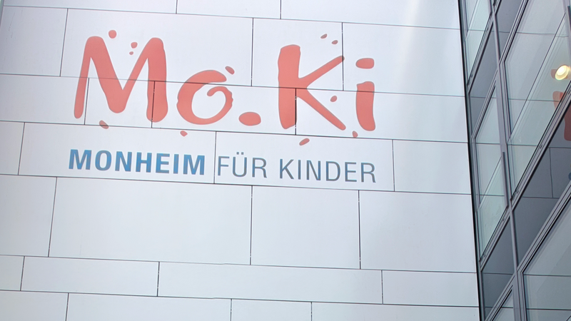 Man sieht man die Fassade eines modernen Gebäudes mit dem großen Schriftzug „Mo.Ki – Monheim für Kinder“. Links oben ist ein bewölkter Himmel zu sehen; rechts erkennt man Glasfenster und eine dunkle Klinkerfassade.