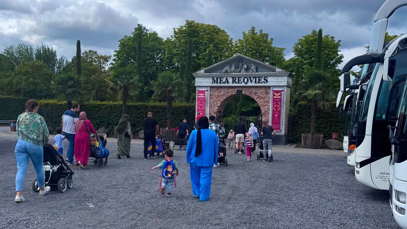 Zu sehen ist der Eingang zum Freizeitpark Irrland mit dem Schriftzug „Mea Requies“ über einem Torbogen. Eine Gruppe von Erwachsenen und Kindern, teils mit Kinderwagen, geht in Richtung Eingang; rechts stehen Reisebusse. Der Himmel ist stark bewölkt, und das Gelände davor besteht aus einer breiten Kiesfläche. Palmen, hohe Zypressen und die klassizistisch wirkende Toranlage verleihen der Szene ein mediterranes Erscheinungsbild.