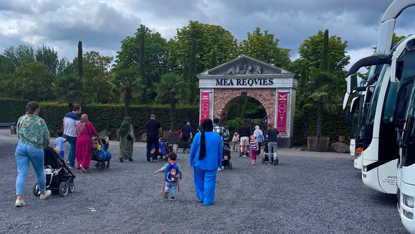 Zu sehen ist der Eingang zum Freizeitpark Irrland mit dem Schriftzug „Mea Requies“ über einem Torbogen. Eine Gruppe von Erwachsenen und Kindern, teils mit Kinderwagen, geht in Richtung Eingang; rechts stehen Reisebusse. Der Himmel ist stark bewölkt, und das Gelände davor besteht aus einer breiten Kiesfläche. Palmen, hohe Zypressen und die klassizistisch wirkende Toranlage verleihen der Szene ein mediterranes Erscheinungsbild.