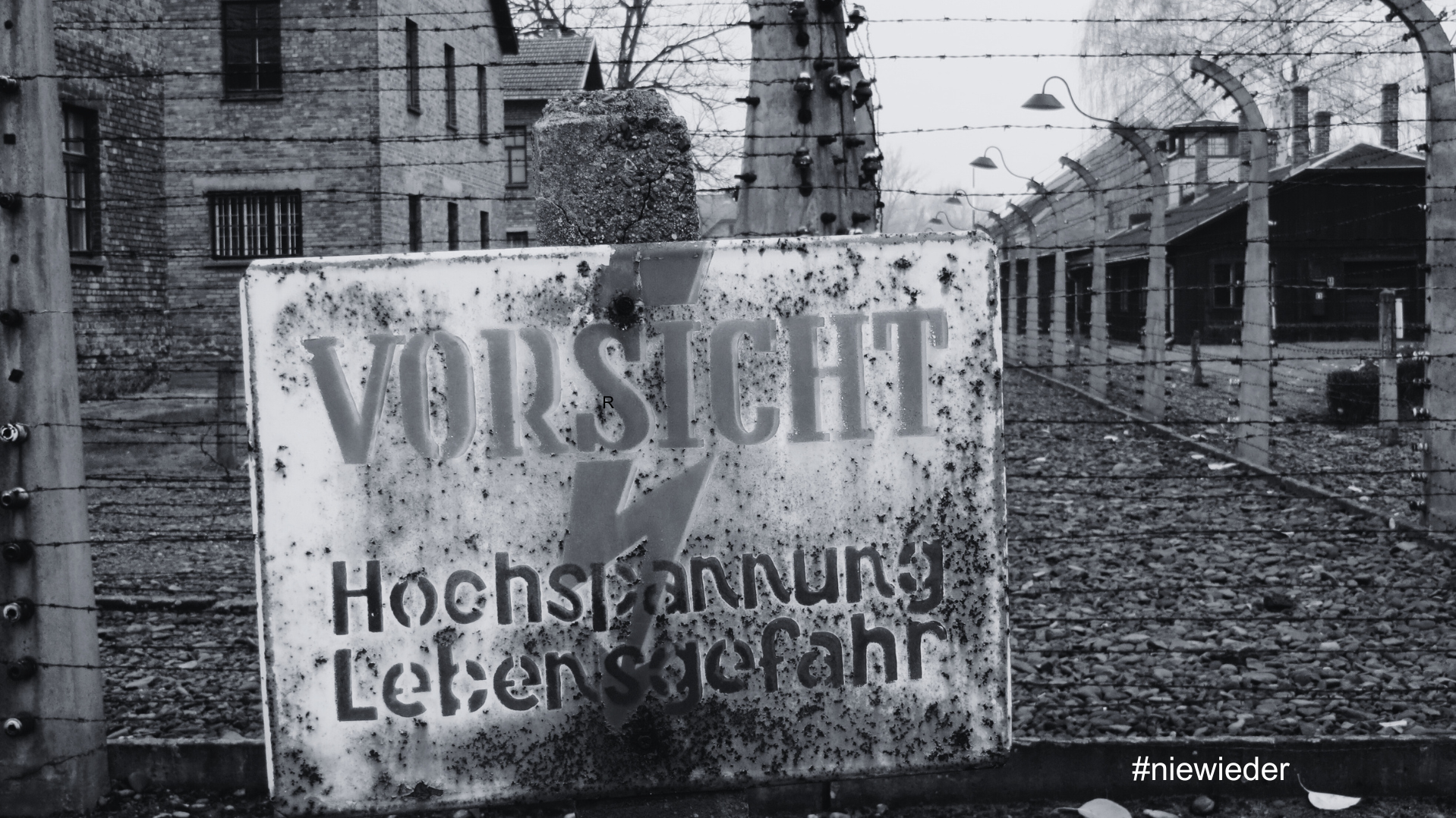 Auf dem Bild ist ein schwarz‑weiß aufgenommenes Konzentrationslager mit Stacheldrahtzäunen und Baracken im Hintergrund zu sehen, im Vordergrund steht ein verwittertes Warnschild mit der Aufschrift „VORSICHT Hochspannung Lebensgefahr“. Bildinhalt Zentral im Bild befindet sich ein rechteckiges, stark verrostetes Schild mit der großen Aufschrift „VORSICHT“ und darunter „Hochspannung Lebensgefahr“, dazu ein Blitzsymbol als Warnzeichen. Hinter dem Schild verlaufen mehrere Reihen Stacheldrahtzaun mit Isolatoren an Betonpfosten, dahinter stehen Backstein- und Holzbaracken, wie sie aus historischen Fotografien nationalsozialistischer Konzentrations- und Vernichtungslager bekannt sind. Kontext und Botschaft Die Perspektive lenkt den Blick durch den Zaun in das Lagerinnere und vermittelt Enge, Bedrohung und das Gefühl von Gefangenschaft, was an die systematische Entrechtung und Ermordung von Menschen im Nationalsozialismus erinnert. Unten rechts ist der Hashtag „#niewieder“ zu lesen, der das Bild klar als Mahnung versteht und zum Erinnern an die Verbrechen und zur Verantwortung gegen Antisemitismus, Rassismus und Menschenfeindlichkeit aufruft.
