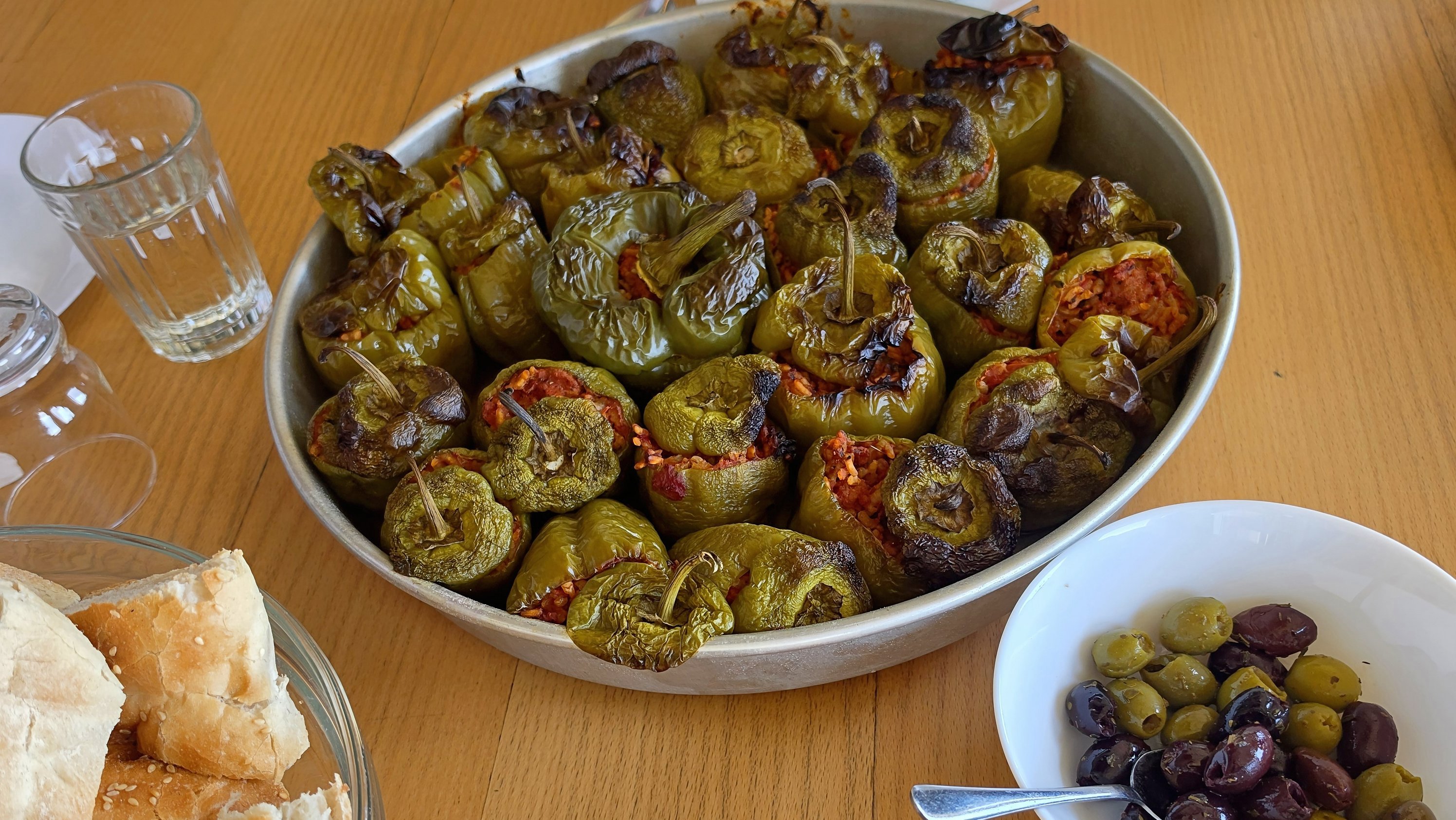 Auf dem Foto ist ein gedeckter Esstisch mit einem griechisch wirkenden Essen zu sehen. In der Mitte steht eine große runde Form mit vielen gebackenen, gefüllten Paprikaschoten, die oben gebräunt sind – wahrscheinlich mit Reis und/oder Hackfleisch gefüllt. Links unten sieht man in einer Schale Stücke von Fladenbrot, rechts unten in einer kleinen weißen Schale gemischte Oliven mit einem Löffel. Im Hintergrund stehen Gläser, Teller mit Besteck und eine Blumendekoration