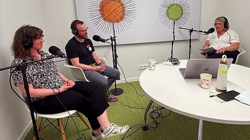 Auf dem Foto sind drei Personen zu sehen, die in einem Raum einen Podcast aufnehmen. In der Mitte steht ein runder, weißer Tisch mit einem Laptop und einem Mikrofon. Zwei der Personen tragen Kopfhörer und sitzen auf weißen Stühlen vor Mikrofonen. Eine dritte Person sitzt ebenfalls mit Kopfhörern und einem Mikrofon am Tisch. An der Wand hinter ihnen hängt ein großes Bild mit zwei stilisierten, sonnenähnlichen Motiven in Orange und Grün. Der Boden ist mit einem grünen Teppich ausgelegt.