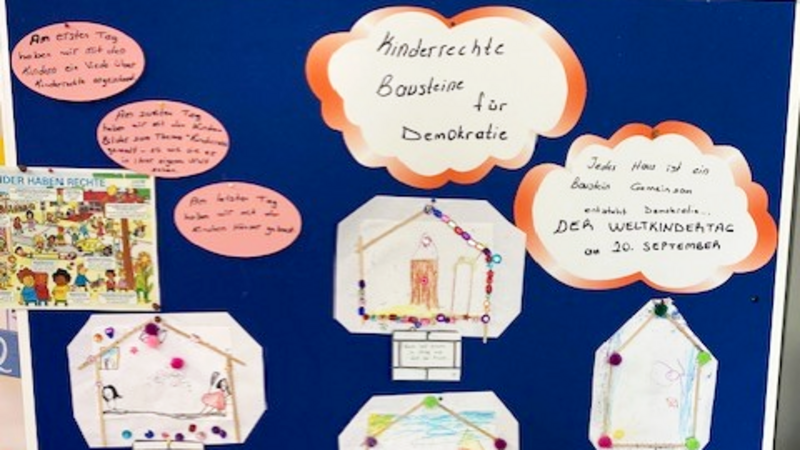 Auf dem Foto ist eine blaue Pinnwand zu sehen, an der mehrere Bilder befestigt sind. Die Bilder sind Teil eines Projekts zum Thema "Kinderrechte – Bausteine für Demokratie", wie die große Überschrift in der Mitte der Pinnwand zeigt. Die Pinnwand ist mit verschiedenen Elementen geschmückt: In der oberen Mitte befindet sich die Überschrift "Kinderrechte Bausteine für Demokratie" in einer wolkenförmigen Sprechblase. Daneben, ebenfalls in einer Sprechblase, steht "Jedes Haus ist ein Baustein Gemeinschaft" und "DER WELTKINDERTAG 20. September". An der linken Seite sind handschriftliche Notizen angebracht, die beschreiben, was die Kinder an verschiedenen Tagen gelernt haben, zum Beispiel "Am ersten Tag haben wir ein Lied über Kinderrechte angeschaut..." und "Am zweiten Tag sind wir ein Rollenspiel zum Thema...". Der größte Teil der Pinnwand ist mit zahlreichen, von Kindern gezeichneten Bildern gefüllt. Jedes dieser Bilder ist in der Form eines Hauses dargestellt und scheint einen Aspekt der Kinderrechte zu illustrieren. Die Häuser sind kreativ gestaltet und teilweise mit kleinen Perlen verziert. Das Foto scheint eine Ausstellung oder eine Projektarbeit aus einer Schule oder einem Kindergarten zu zeigen, die im Rahmen des Weltkindertags am 20. September entstanden ist.