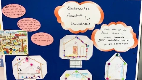 Auf dem Foto ist eine blaue Pinnwand zu sehen, an der mehrere Bilder befestigt sind. Die Bilder sind Teil eines Projekts zum Thema "Kinderrechte – Bausteine für Demokratie", wie die große Überschrift in der Mitte der Pinnwand zeigt. Die Pinnwand ist mit verschiedenen Elementen geschmückt: In der oberen Mitte befindet sich die Überschrift "Kinderrechte Bausteine für Demokratie" in einer wolkenförmigen Sprechblase. Daneben, ebenfalls in einer Sprechblase, steht "Jedes Haus ist ein Baustein Gemeinschaft" und "DER WELTKINDERTAG 20. September". An der linken Seite sind handschriftliche Notizen angebracht, die beschreiben, was die Kinder an verschiedenen Tagen gelernt haben, zum Beispiel "Am ersten Tag haben wir ein Lied über Kinderrechte angeschaut..." und "Am zweiten Tag sind wir ein Rollenspiel zum Thema...". Der größte Teil der Pinnwand ist mit zahlreichen, von Kindern gezeichneten Bildern gefüllt. Jedes dieser Bilder ist in der Form eines Hauses dargestellt und scheint einen Aspekt der Kinderrechte zu illustrieren. Die Häuser sind kreativ gestaltet und teilweise mit kleinen Perlen verziert. Das Foto scheint eine Ausstellung oder eine Projektarbeit aus einer Schule oder einem Kindergarten zu zeigen, die im Rahmen des Weltkindertags am 20. September entstanden ist.