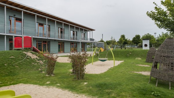 Auf dem Bild ist das Außengelände einer Kindertagesstätte zu sehen. Im Hintergrund steht ein modernes zweigeschossiges Gebäude mit großen Fenstern und einem Balkon. Davor befinden sich: eine große Rasenfläche, ein Sandbereich mit einer Schaukel, eine rote Rutsche, weitere Spielgeräte, ein Weiden- bzw. Kletterhäuschen, Bäume und Sträucher.
