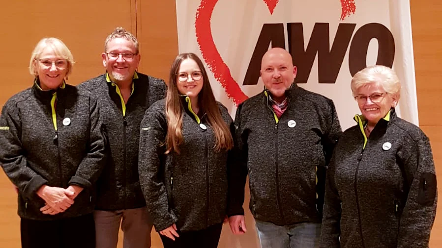 Das Foto zeigt eine Gruppe von fünf Personen, die gemeinsam vor einem Banner mit einem Herzsymbol und der Aufschrift „AWO“ (vermutlich Arbeiterwohlfahrt) stehen. Alle tragen ähnliche Jacken mit grünlichen Akzenten.