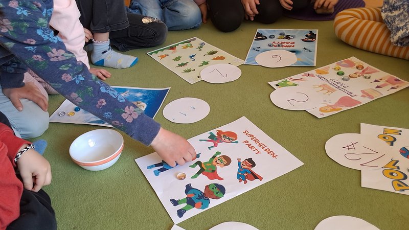 Auf dem Bild sitzen mehrere Kinder im Kreis auf einem grünen Teppichboden. In der Mitte liegen verschiedene bunte Arbeitsblätter und runde Papierkarten mit Zahlen darauf. Die Blätter zeigen thematische Bilder – zum Beispiel Superhelden, Tiere oder andere Figuren. Ein Kind greift gerade nach einem der Blätter mit Superheldenmotiven. Neben den Materialien steht eine kleine Schüssel. Die Kinder wirken konzentriert, als würden sie gemeinsam ein Spiel oder eine Lernaktivität durchführen, bei der Bilder und Zahlen zugeordnet werden.