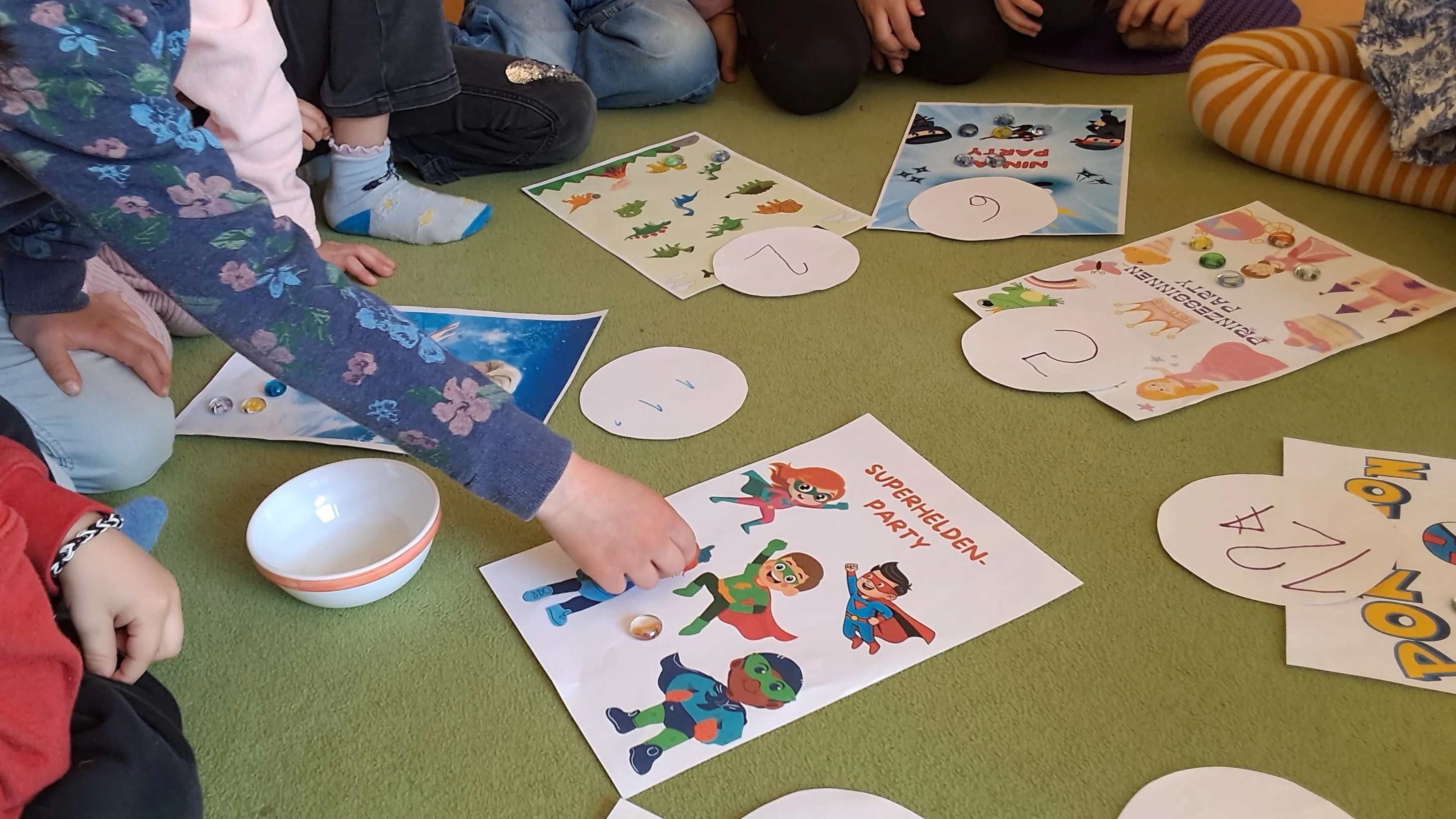 Auf dem Bild sitzen mehrere Kinder im Kreis auf einem grünen Teppichboden. In der Mitte liegen verschiedene bunte Arbeitsblätter und runde Papierkarten mit Zahlen darauf. Die Blätter zeigen thematische Bilder – zum Beispiel Superhelden, Tiere oder andere Figuren. Ein Kind greift gerade nach einem der Blätter mit Superheldenmotiven. Neben den Materialien steht eine kleine Schüssel. Die Kinder wirken konzentriert, als würden sie gemeinsam ein Spiel oder eine Lernaktivität durchführen, bei der Bilder und Zahlen zugeordnet werden.