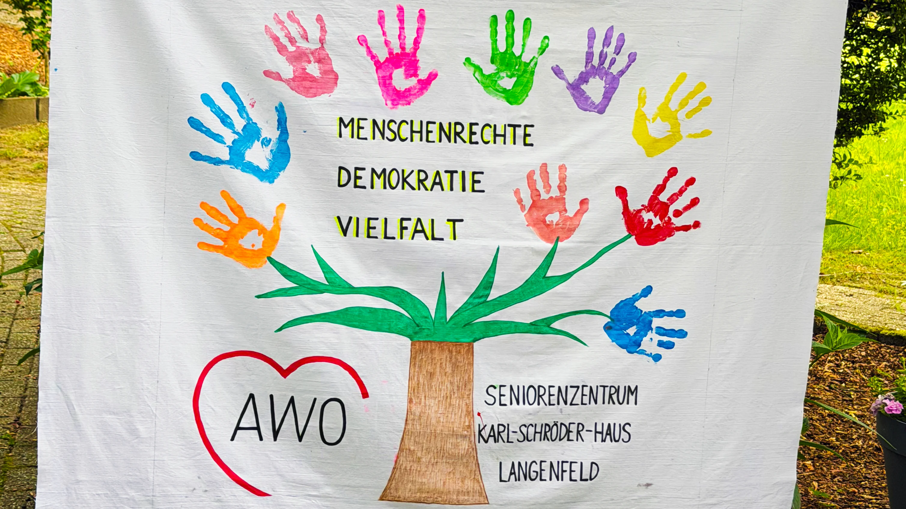 Auf dem Foto ist ein großes, bemaltes Stofftuch zu sehen, das von zwei Personen an den Seiten hochgehalten wird. In der Mitte ist ein Baum gemalt, dessen Krone aus bunten Handabdrücken in verschiedenen Farben besteht, die wie Blätter wirken. Auf dem Tuch stehen die Worte „MENSCHENRECHTE“, „DEMOKRATIE“ und „VIELFALT“ sowie unten links ein rotes Herz mit der Aufschrift „AWO“. Rechts unten steht „SENIORENZENTRUM KARL-SCHRÖDER-HAUS LANGENFELD“.