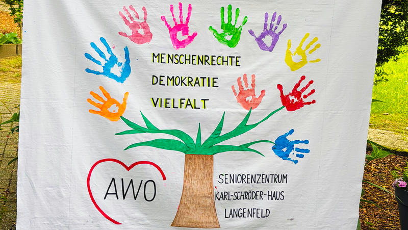Auf dem Foto ist ein großes, bemaltes Stofftuch zu sehen, das von zwei Personen an den Seiten hochgehalten wird. In der Mitte ist ein Baum gemalt, dessen Krone aus bunten Handabdrücken in verschiedenen Farben besteht, die wie Blätter wirken. Auf dem Tuch stehen die Worte „MENSCHENRECHTE“, „DEMOKRATIE“ und „VIELFALT“ sowie unten links ein rotes Herz mit der Aufschrift „AWO“. Rechts unten steht „SENIORENZENTRUM KARL-SCHRÖDER-HAUS LANGENFELD“.