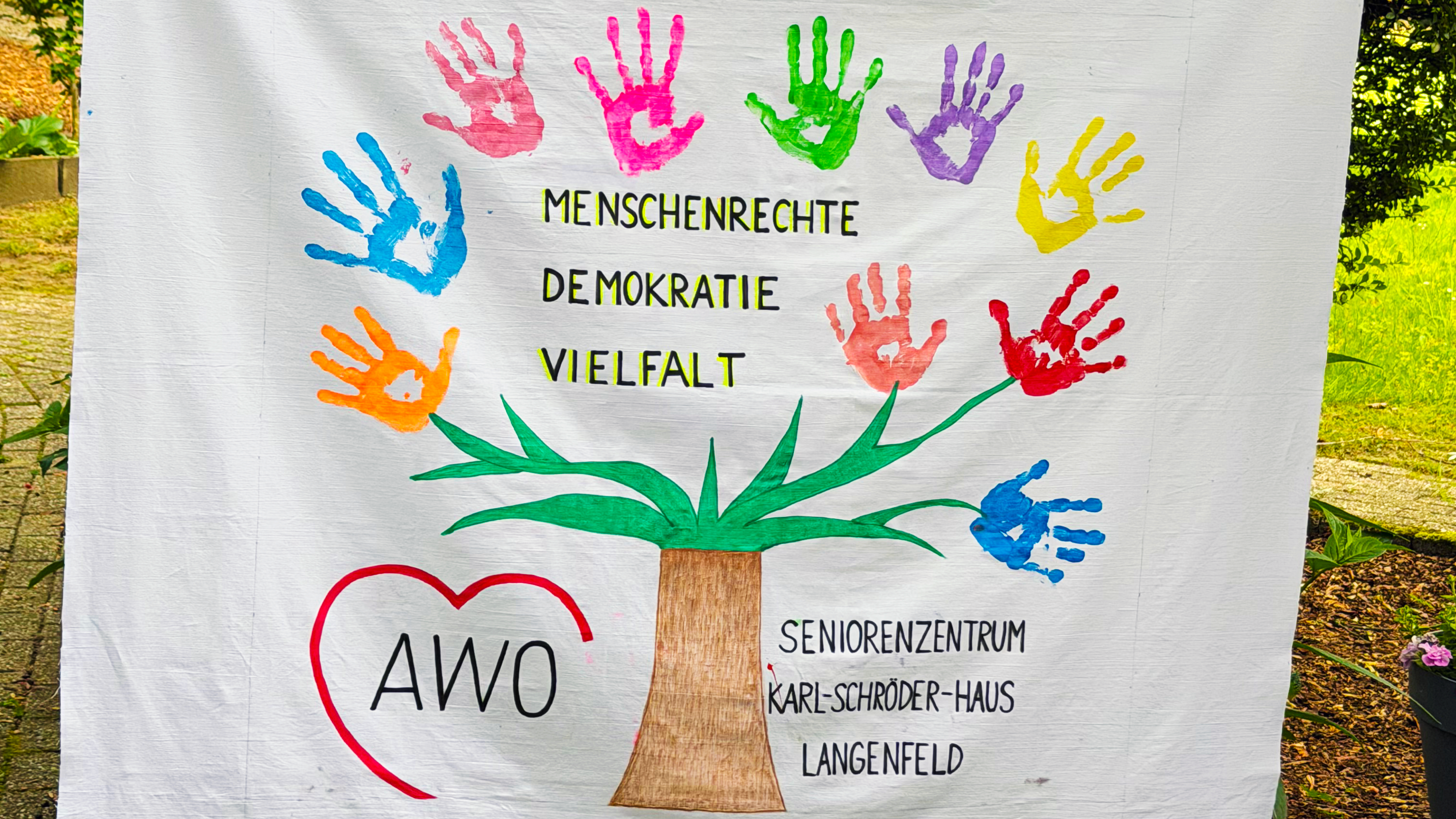 Auf dem Foto ist ein großes, bemaltes Stofftuch zu sehen, das von zwei Personen an den Seiten hochgehalten wird. In der Mitte ist ein Baum gemalt, dessen Krone aus bunten Handabdrücken in verschiedenen Farben besteht, die wie Blätter wirken. Auf dem Tuch stehen die Worte „MENSCHENRECHTE“, „DEMOKRATIE“ und „VIELFALT“ sowie unten links ein rotes Herz mit der Aufschrift „AWO“. Rechts unten steht „SENIORENZENTRUM KARL-SCHRÖDER-HAUS LANGENFELD“.