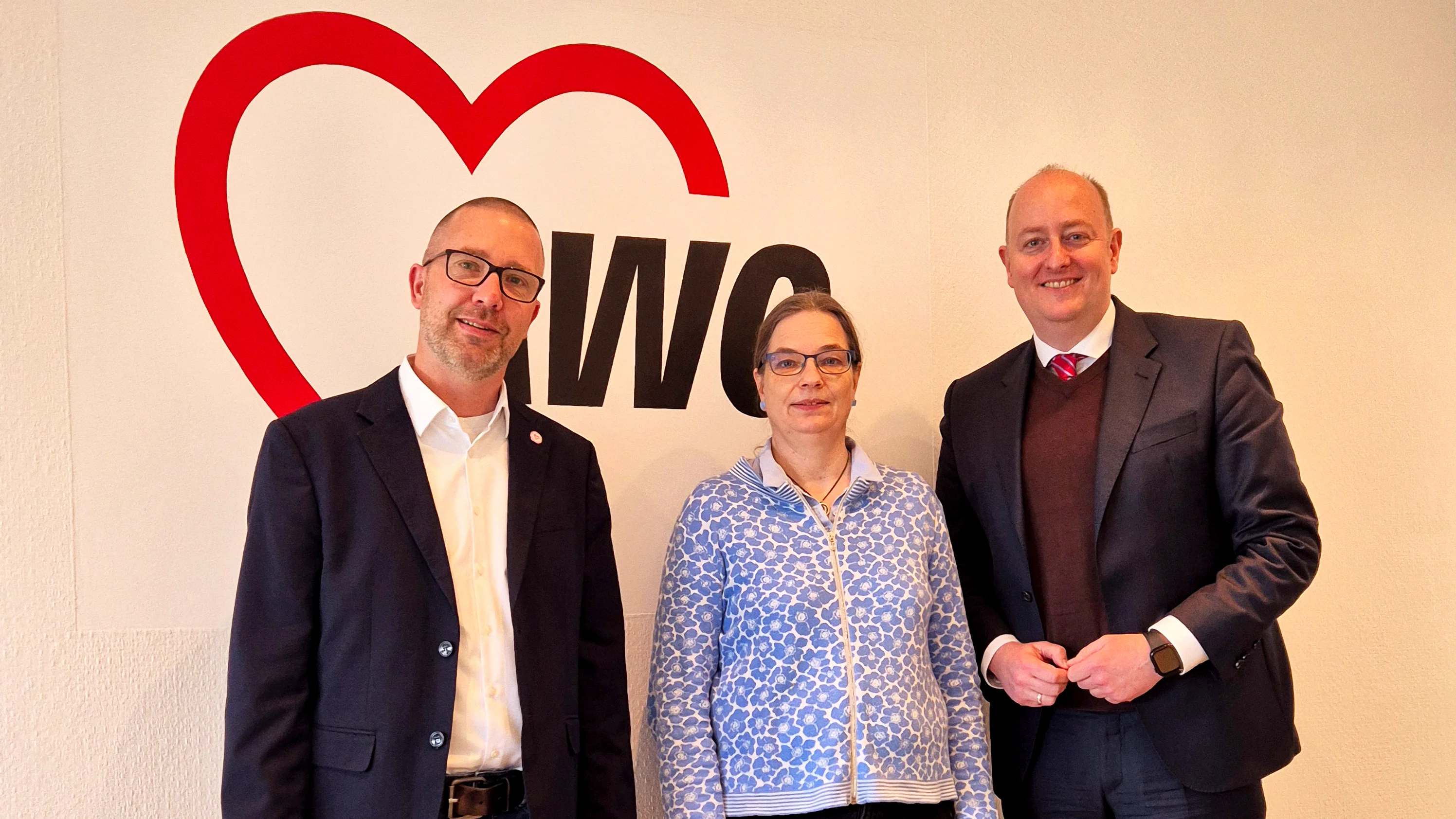 Das Foto zeigt drei Personen, die vor einem Logo mit den Buchstaben "AWO" und einem stilisierten roten Herz stehen. Die Abkürzung "AWO" steht für die Arbeiterwohlfahrt, eine deutsche Wohlfahrtsorganisation. Die drei Personen tragen geschäftliche oder semi-formelle Kleidung und posieren für das Foto.