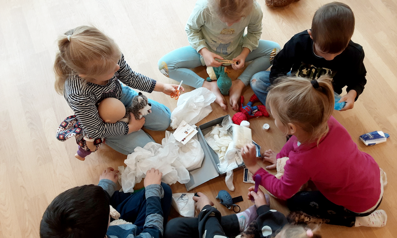 Auf dem Bild sind mehrere kleine Kinder zu sehen, die auf einem hellen Holzboden im Kreis sitzen. Sie beschäftigen sich intensiv mit Stofftieren und verschiedenen Materialien, die wie medizinische Utensilien wirken – darunter: Ein Karton mit weißen Handschuhen, Taschentücher oder Mulltücher, Spielzeug-Spritzen, Scheren (vermutlich Kinderscheren), Stofftiere wie ein kleiner Wolf, ein Dinosaurier, ein Vogel, ein Teddybär und eine Spiderman-Figur. Es sieht so aus, als würden die Kinder einen Teddybär-Krankenhaus-Kurs („Teddykurs“) machen, bei dem Kinder spielerisch medizinische Behandlungen nachahmen und Ängste vor Arztbesuchen abbauen können. Dabei „behandeln“ sie ihre Kuscheltiere, was oft in Kindergärten oder Grundschulen durchgeführt wird. Das Ganze wirkt sehr konzentriert und fantasievoll – eine gute Mischung aus Spiel und Lernen.