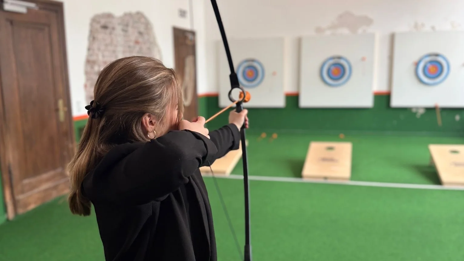 Auf dem Foto ist eine Person in einer Indoor‑Bogenschießanlage zu sehen, die mit einem Bogen auf eine Zielscheibe schießt. Im Hintergrund erkennt man mehrere Zielscheiben an der Wand sowie einen Boden mit grünem Kunstrasen und Holz­türen an der Seite.