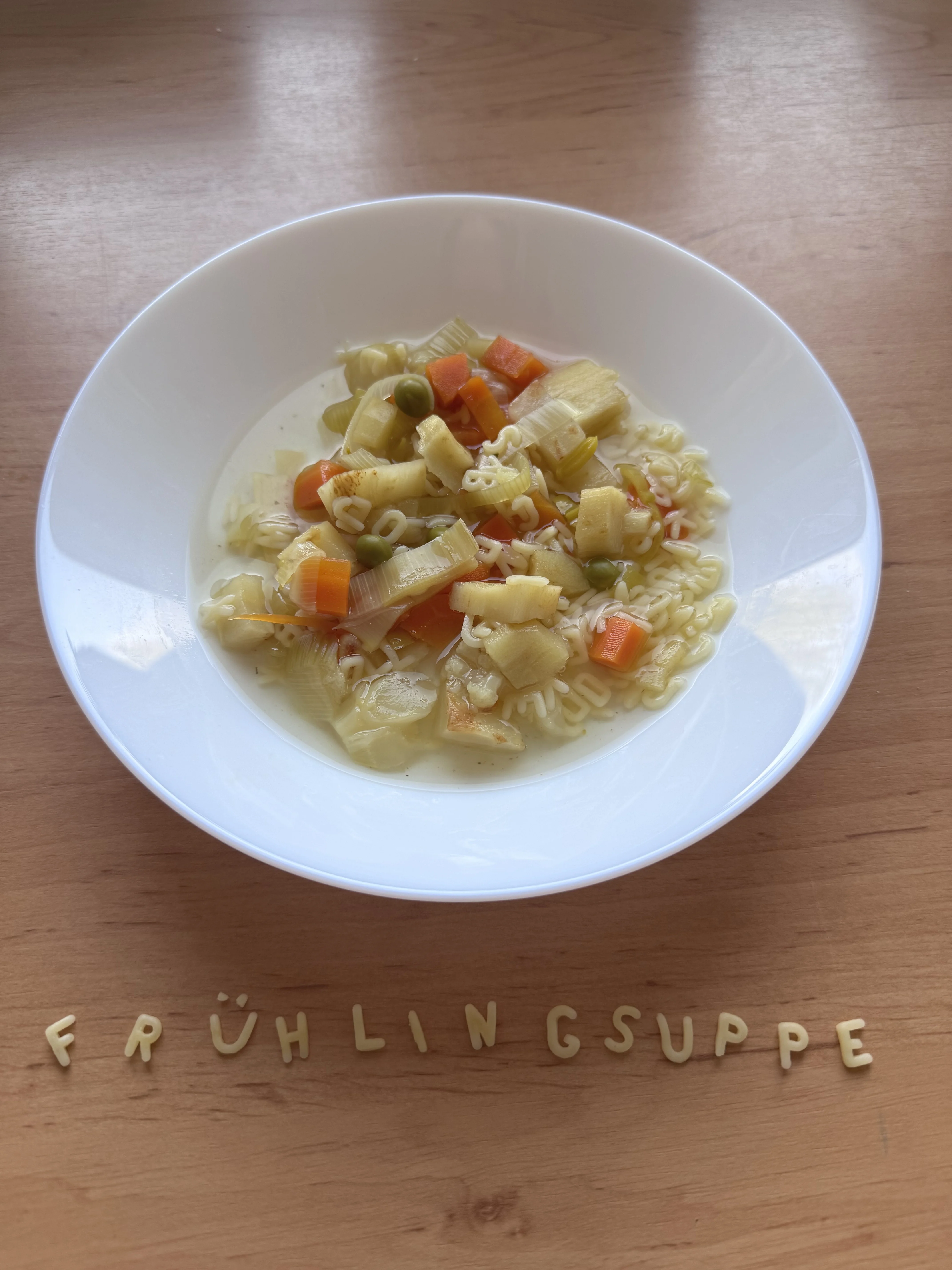 Auf dem Foto steht ein weißer Teller mit einer klaren Gemüsesuppe auf einem Holztisch. In der Suppe sind bunte Gemüsewürfel (z.B. Karotten, Erbsen, Lauch) und Buchstabennudeln zu sehen, und vor dem Teller sind aus Buchstabennudeln die Wörter „FRÜHLING SUPPE“ gelegt.