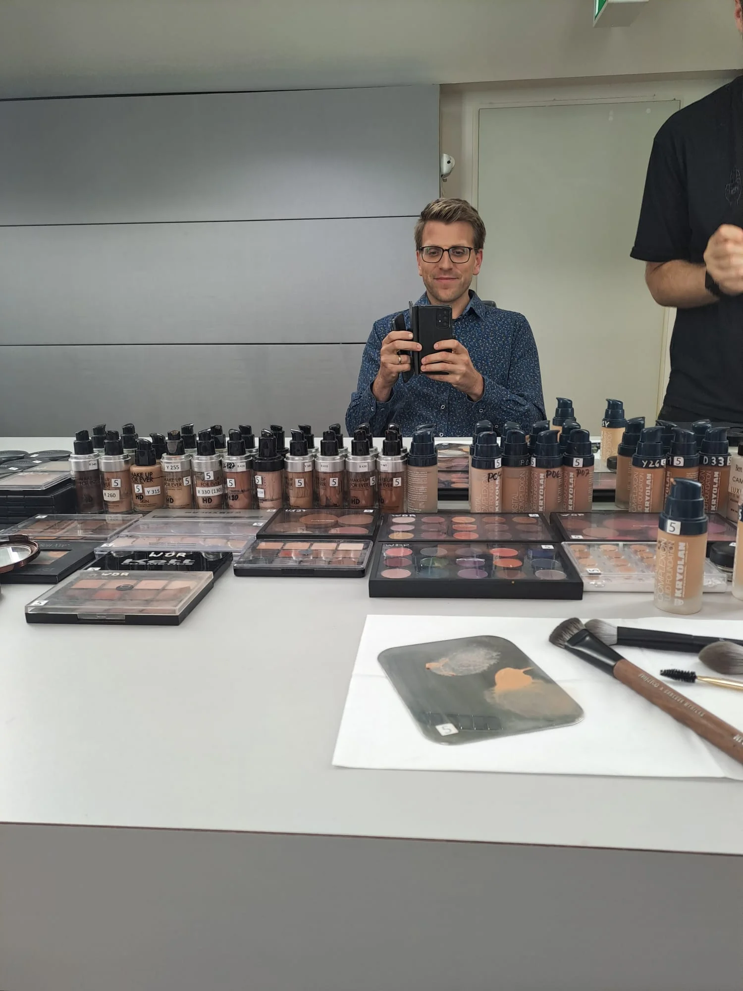 In dem Bild ist eine Person vor einem Spiegel zu sehen, die gerade ein Foto macht.. Auf der Theke vor der Person befinden sich verschiedene Make-up-Produkte wie Flaschen, Paletten und Pinsel.