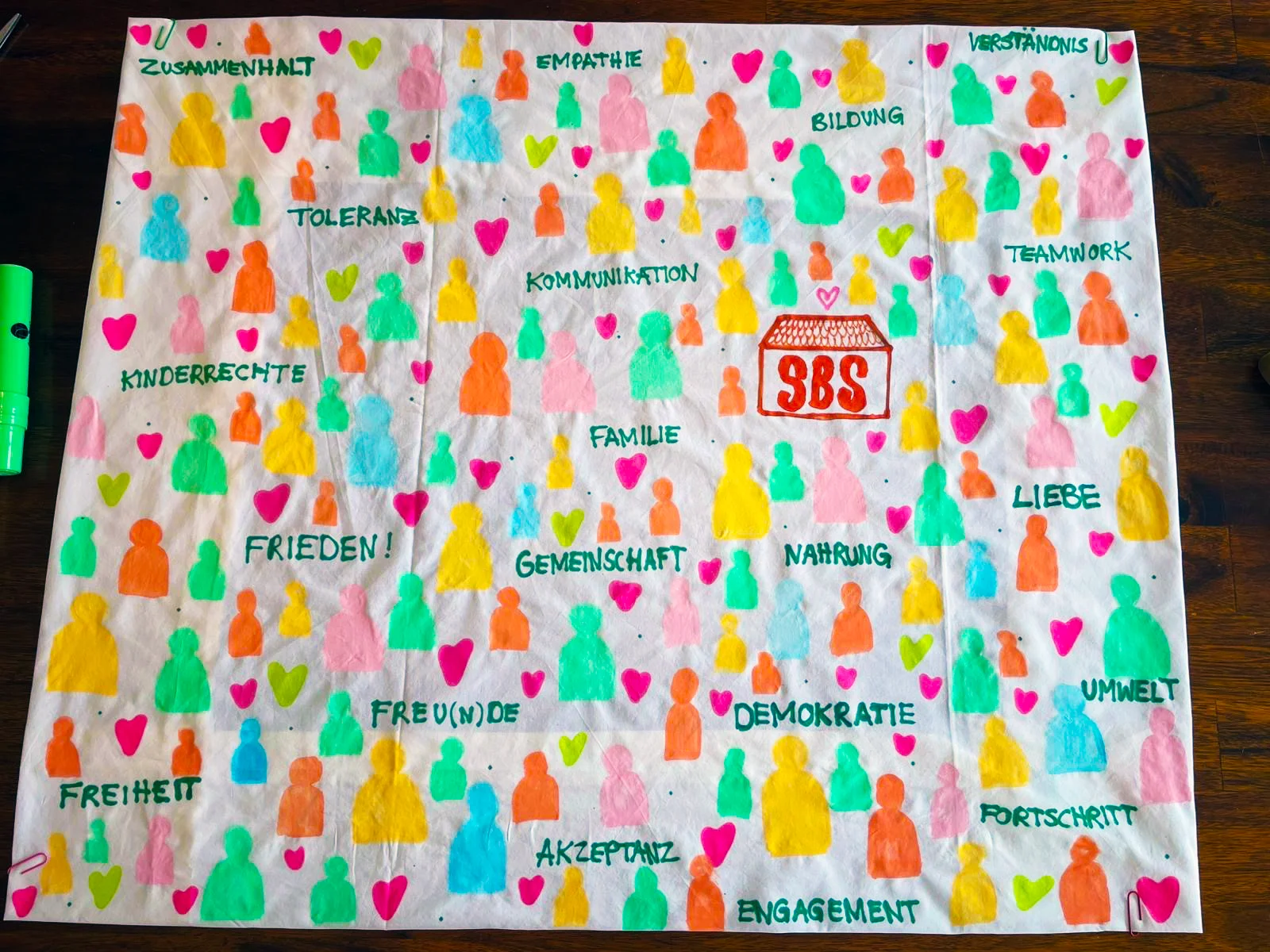 uf dem Bild ist ein bemaltes Stofftuch zu sehen, das dicht mit bunten Silhouetten von Menschen und vielen farbigen Herzen gestaltet ist. Zwischen den Figuren stehen mit Filzstift Begriffe wie „ZUSAMMENHALT“, „EMPATHIE“, „VERSTÄNDNIS“, „TOLERANZ“, „BILDUNG“, „TEAMWORK“, „KINDERRECHTE“, „FAMILIE“, „KOMMUNIKATION“, „FRIEDEN!“, „GEMEINSCHAFT“, „NAHRUNG“, „LIEBE“, „FREIHEIT“, „FREU(N)DE“, „DEMOKRATIE“, „UMWELT“, „FORTSCHRITT“, „AKZEPTANZ“ und „ENGAGEMENT“. In der Mitte ist ein kleines Haus mit dem Schriftzug „SBS“ in Rot gezeichnet.