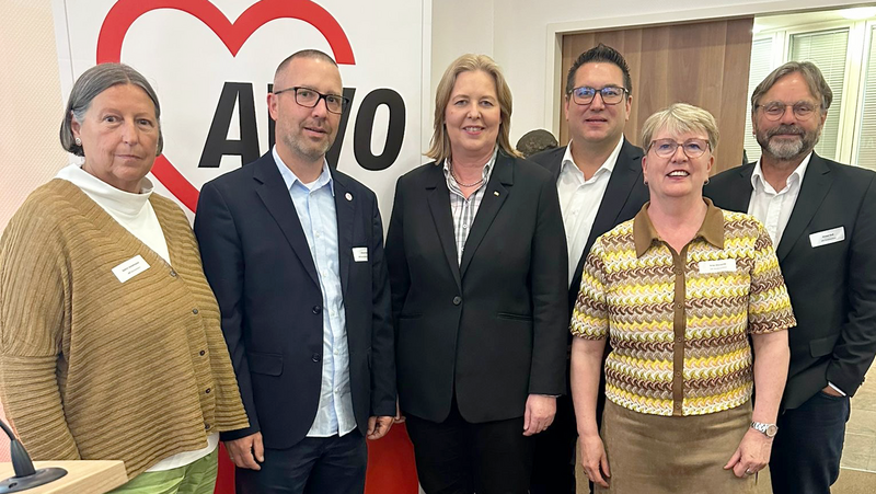 Auf dem Foto sind sechs Personen zu sehen, die gemeinsam vor einem Roll-Up mit dem Logo der AWO (Arbeiterwohlfahrt) stehen. Das AWO-Logo zeigt ein rotes Herz mit dem Schriftzug „AWO“ darin. Die Personen wirken formell oder semi-formell gekleidet. Die Personen tragen Namensschilder, was weiter auf einen offiziellen oder öffentlichen Anlass hindeutet. Der Hintergrund zeigt einen Konferenzraum oder Bürobereich.